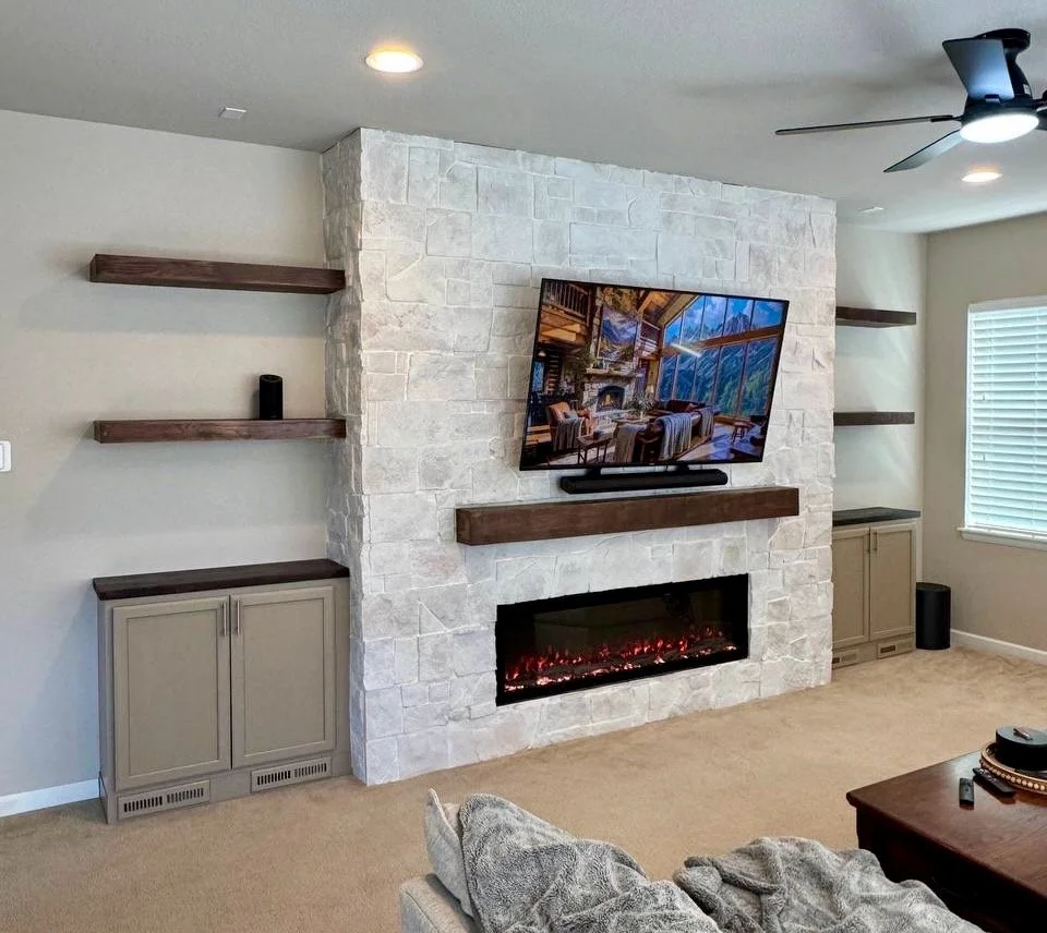 Wide view of a Castle Rock living room with a floor-to-ceiling stone fireplace media wall, symmetrical cabinets, wood shelves, mounted television, and electric fireplace.