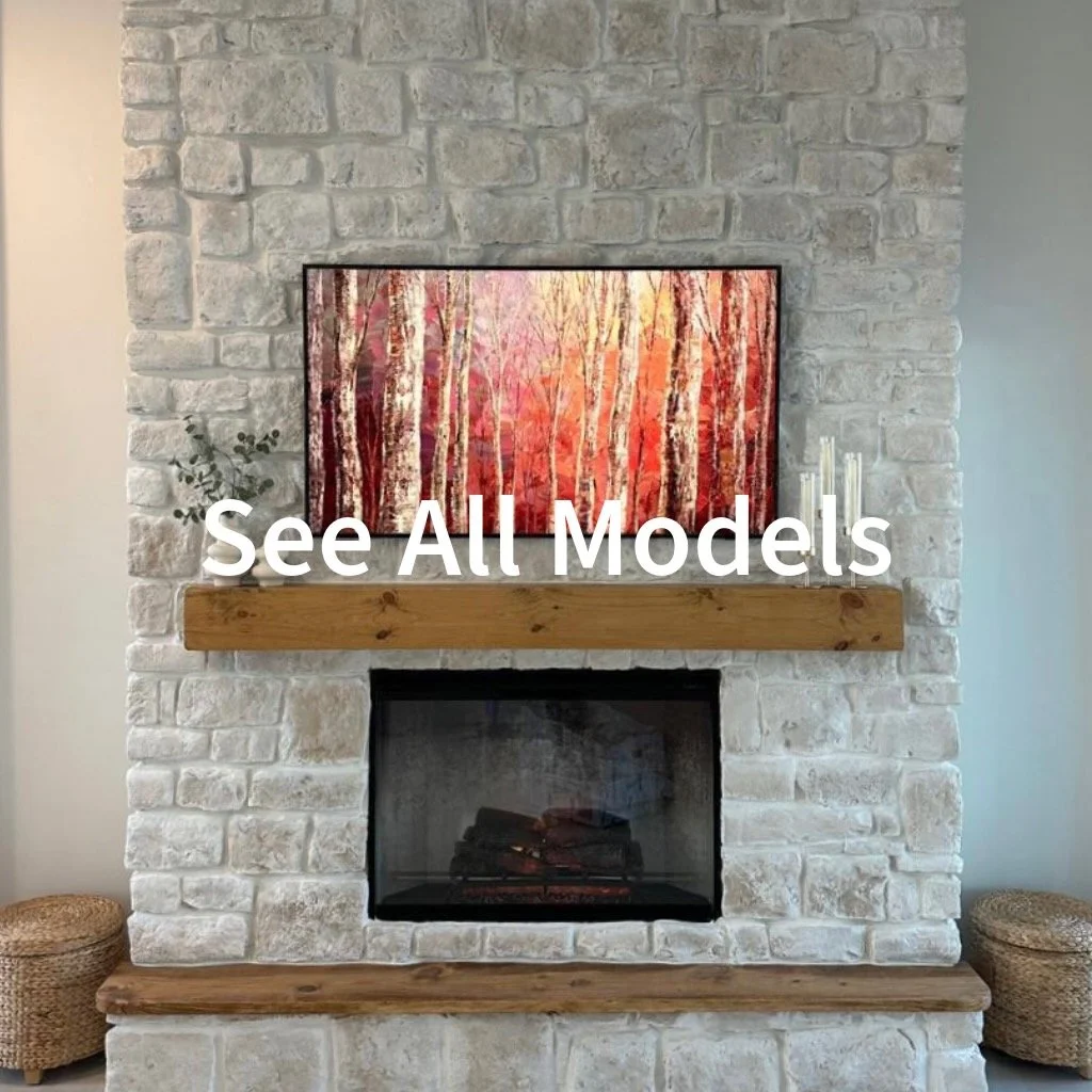 A white brick fireplace with a wooden mantel, a colorful painting of autumn trees above it, and small decorative objects including a plant, white candles, and woven baskets on the floor.