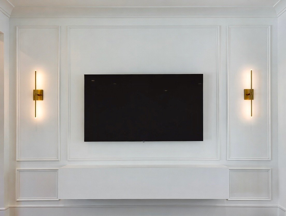 Living room wall with white paneling, mounted flat-screen TV, and two gold wall sconces emitting soft light.
