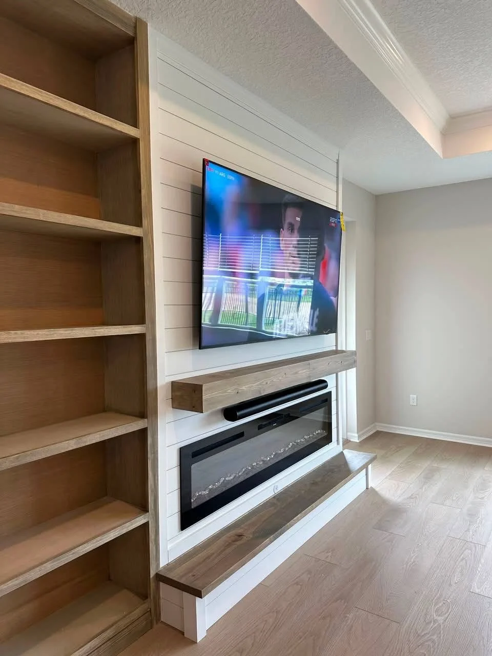 Side perspective of a shiplap media wall with floating wood mantel, electric fireplace, and custom shelving