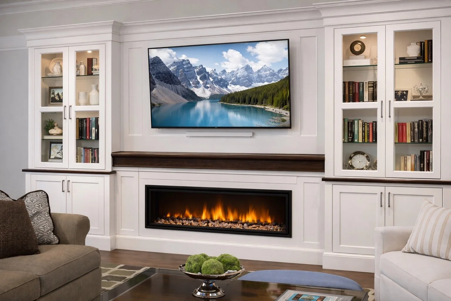 Living room with white built-in cabinets, a wall-mounted TV showing mountain and lake scenery, a fireplace with flames, a beige sofa with pillows, a white armchair with a pillow, a glass table with green decorative spheres, a small blue rug, and a ma
