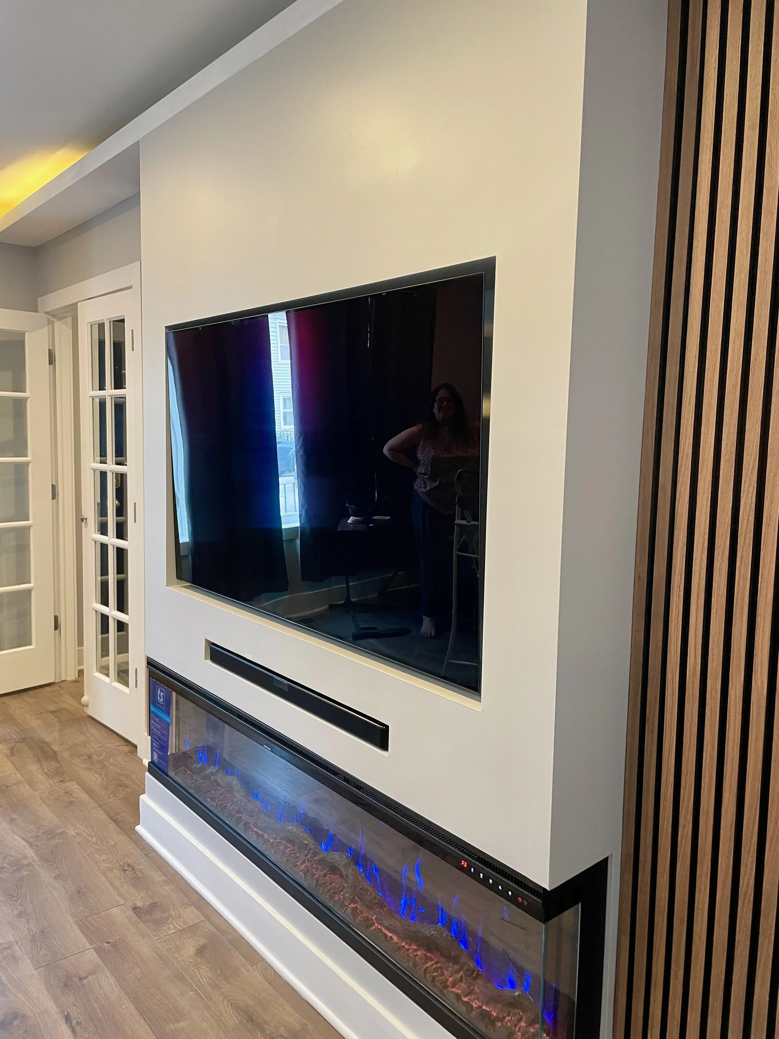 Flush-mounted TV and soundbar on a clean white wall, featuring a long three-sided electric fireplace and wood slat panels on both sides.