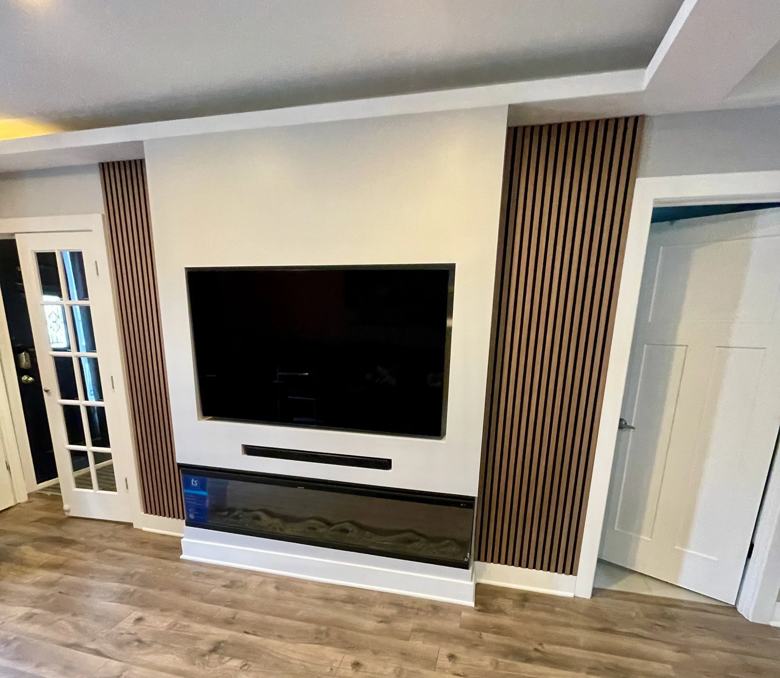 Living room with a wall-mounted television, modern wooden panel accents, a door with glass panes, and wood flooring.