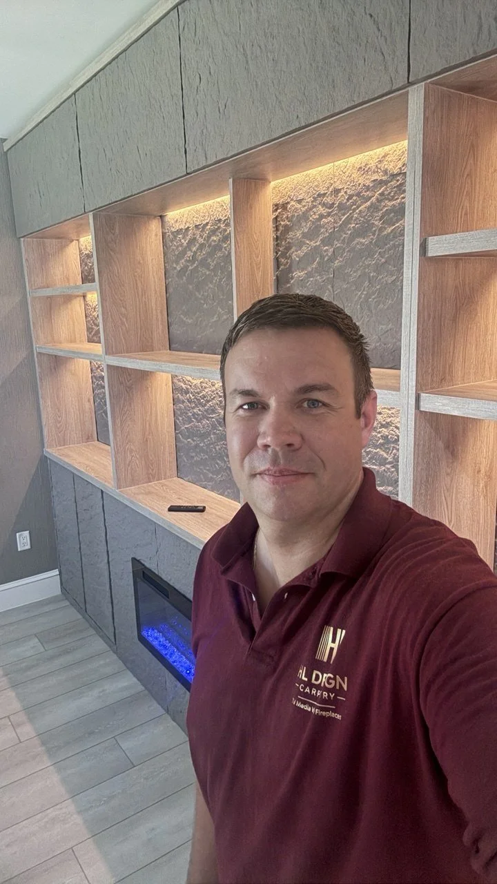 A man taking a selfie in front of a modern interior with built-in wooden shelves, a textured stone wall, and an electric fireplace with blue flames.