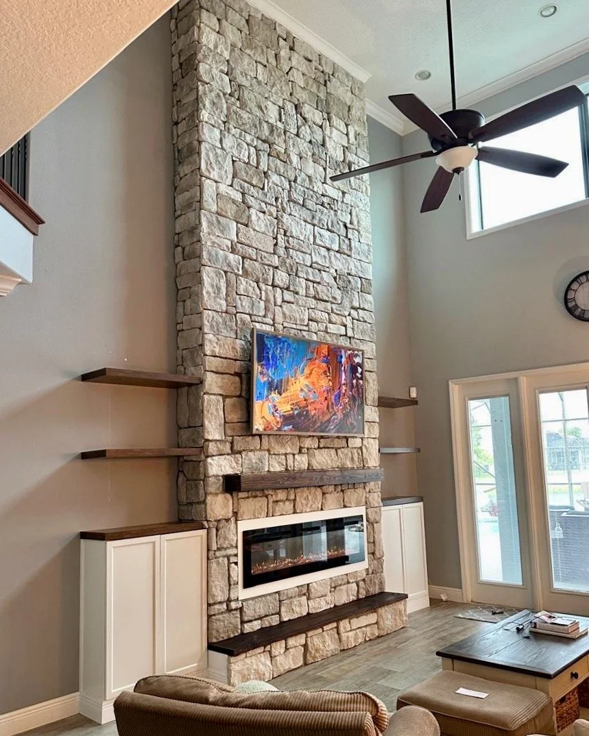 Living room with a tall stone fireplace, floating shelves on either side, a mounted flat-screen TV, a ceiling fan, a clock on the wall, and glass doors leading outside.