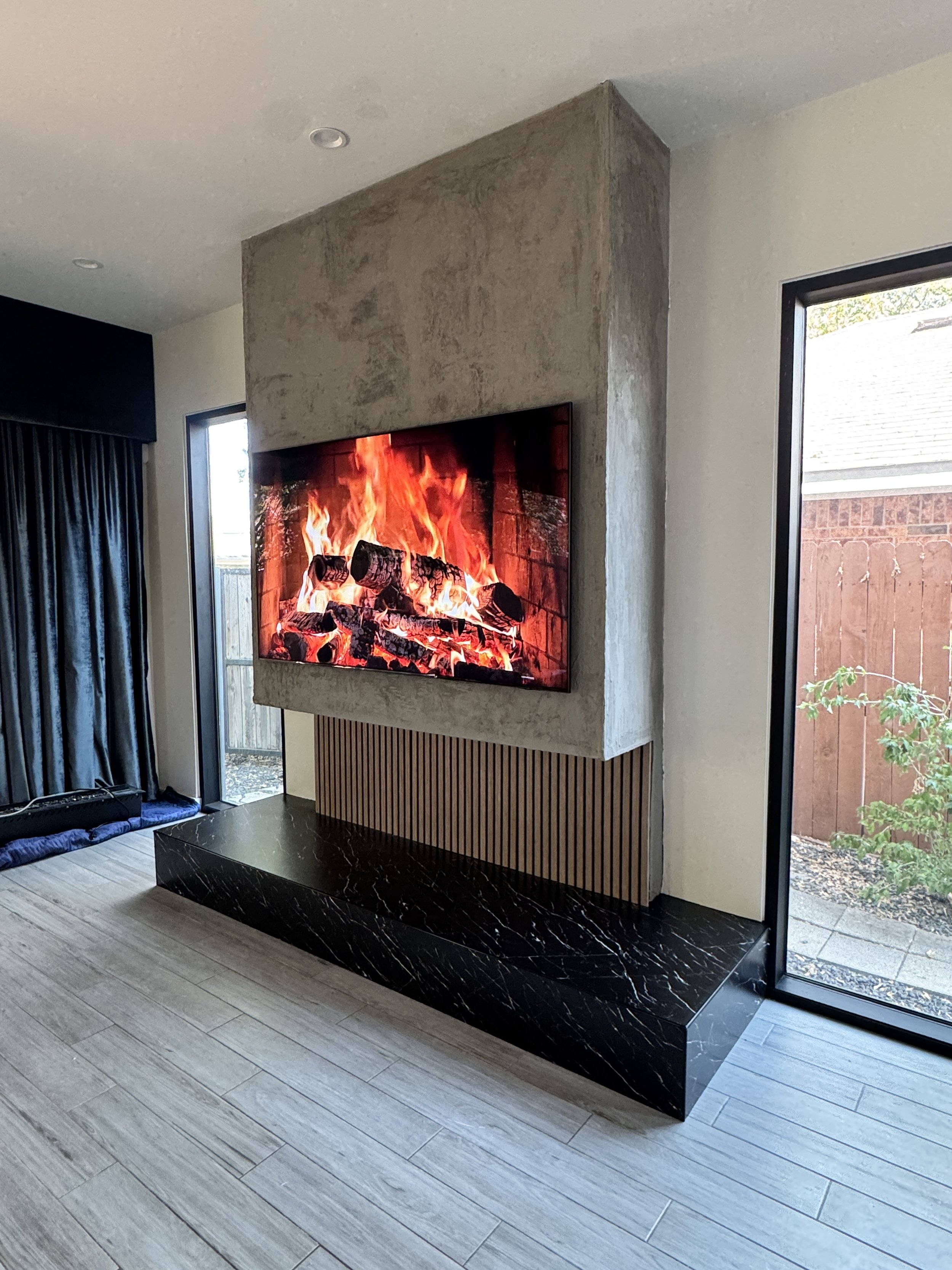 Angled view of a bespoke Dallas media wall showing the projecting concrete-effect TV volume above recessed wood slats and a polished black granite platform.