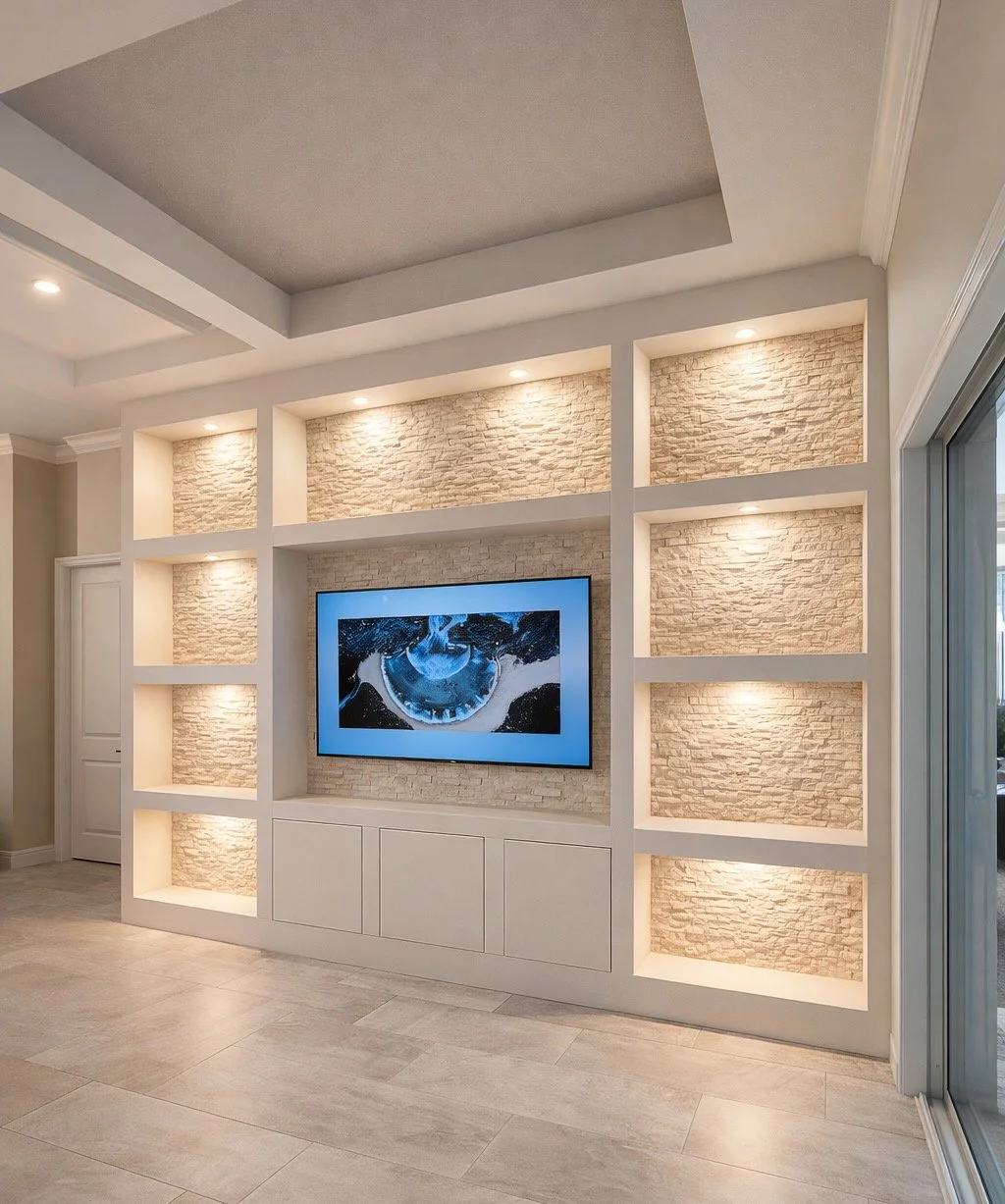 Modern living room wall with built-in shelves illuminated by recessed lighting, a mounted flat-screen TV, and a brick accent wall.