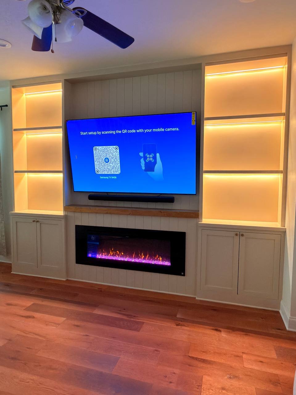 Angled view of an Orlando entertainment built-in with shaker cabinets, warm LED shelf lighting, TV, soundbar, and electric fireplace.