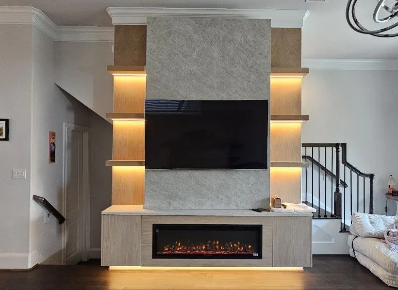 Custom built-in fireplace media wall in Houston with a large TV, stone-look center panel, wood veneer niches, floating cabinet, and warm LED lighting.