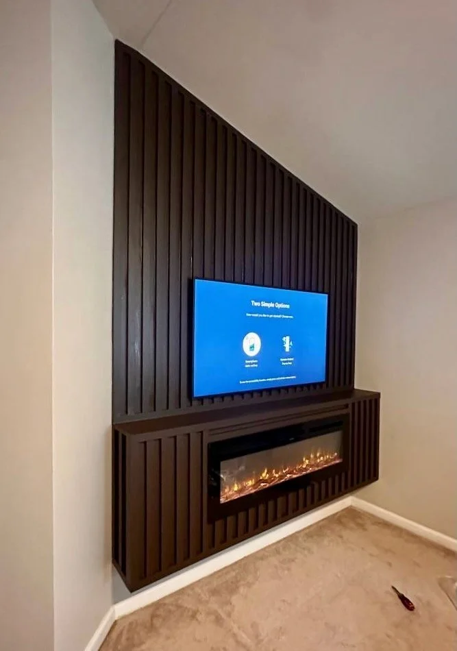 A sleek corner-mounted media wall in a bedroom, featuring a TV above a linear electric fireplace, finished with dark vertical wood slats from floor to ceiling.