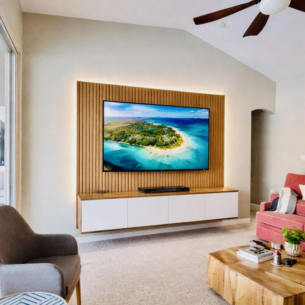 Living room with a wall-mounted flat-screen TV showing a tropical beach scene, wooden panel backdrop, white floating cabinet below, and furniture including a gray armchair, pink sofa, and wooden coffee table.