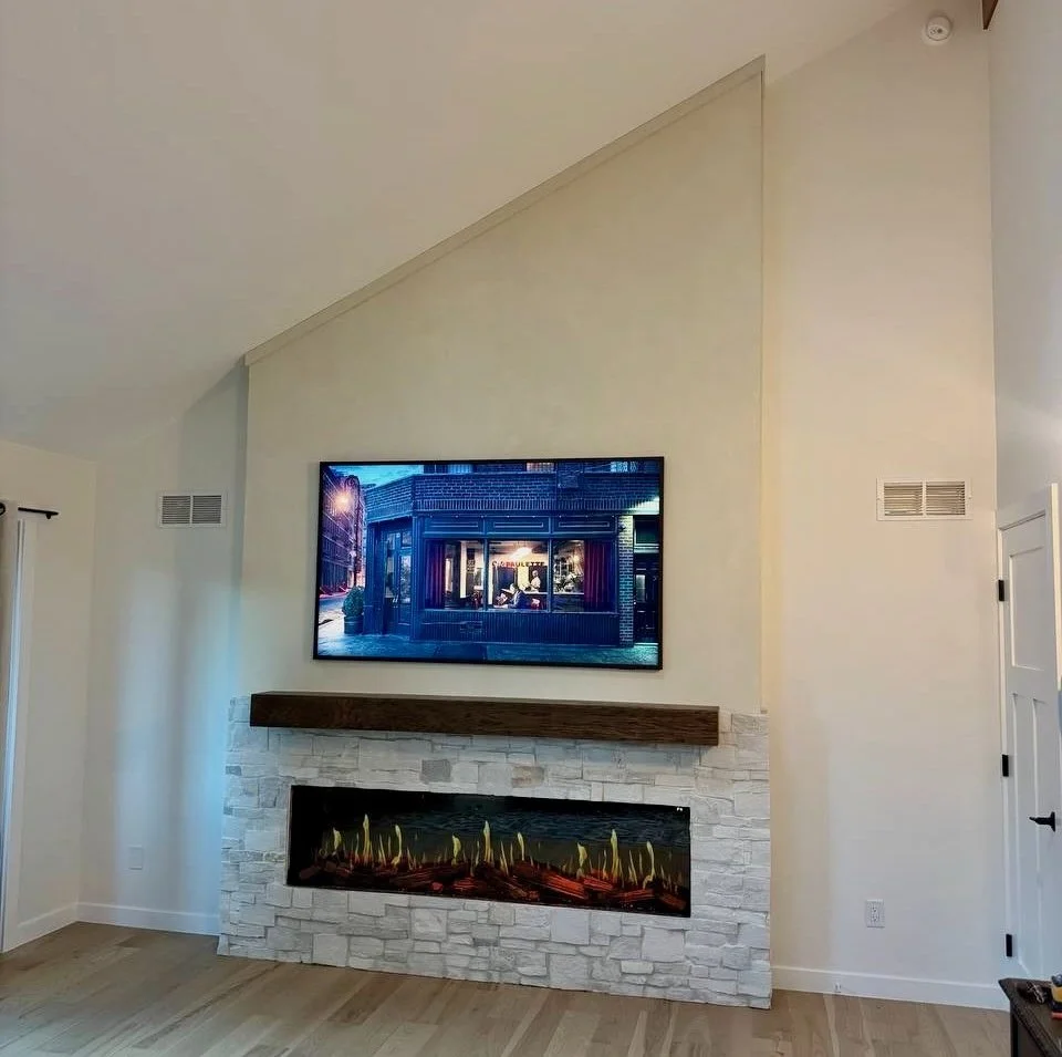 Wide view of a Boulder living room featuring a stone and limewash TV wall, dark wood mantel, electric fireplace, and angled ceiling.
