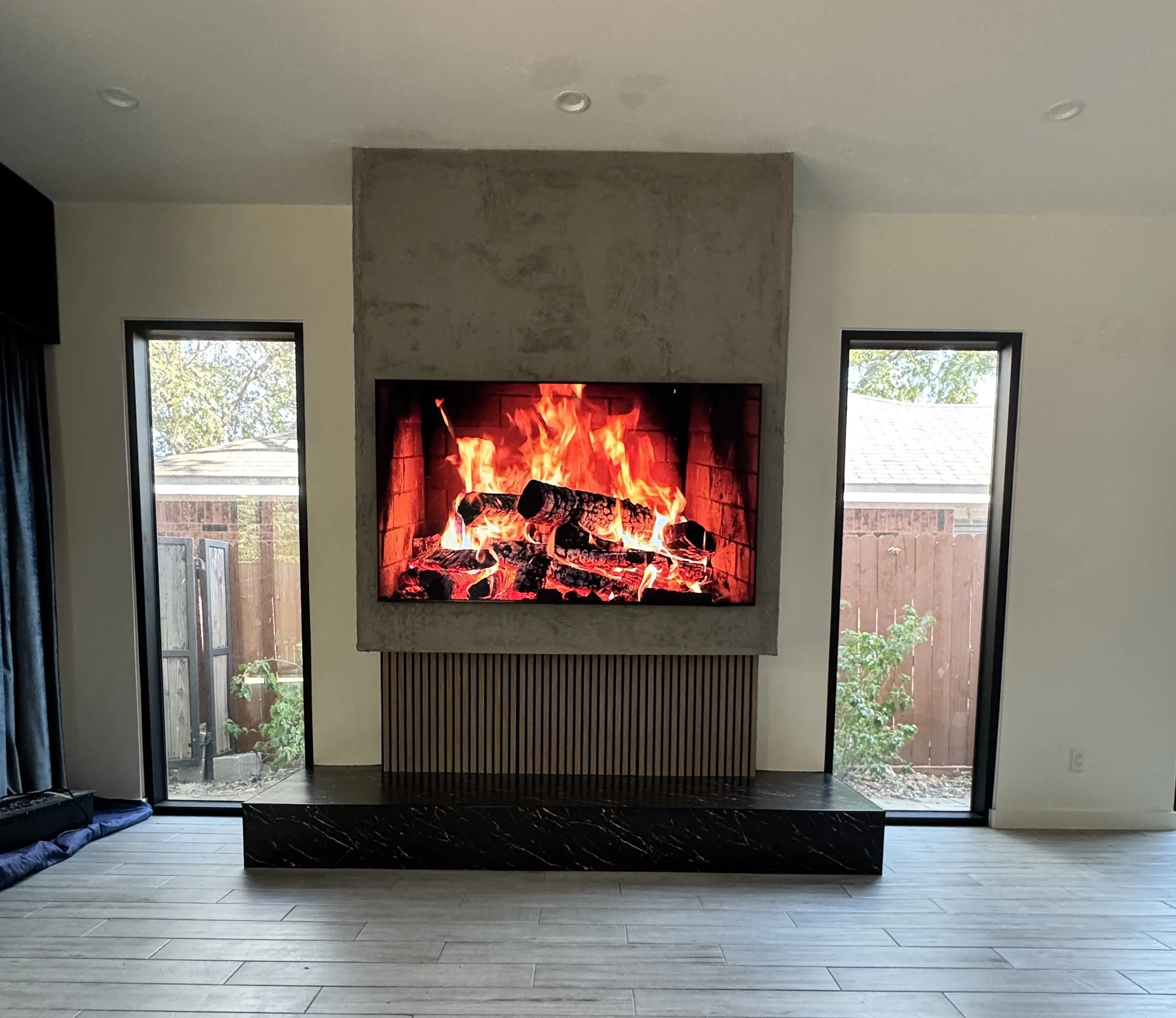 Front view of a custom three-material TV wall centered between narrow windows with a textured concrete finish, wood slats, and a black stone base.