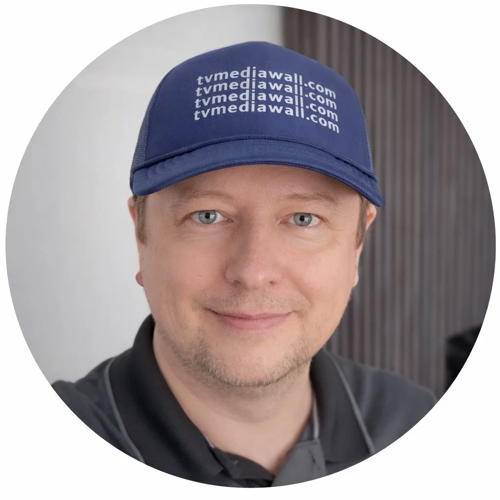 A man wearing a blue cap with 'tvmediawall.com' logo, smiling, against a plain background.