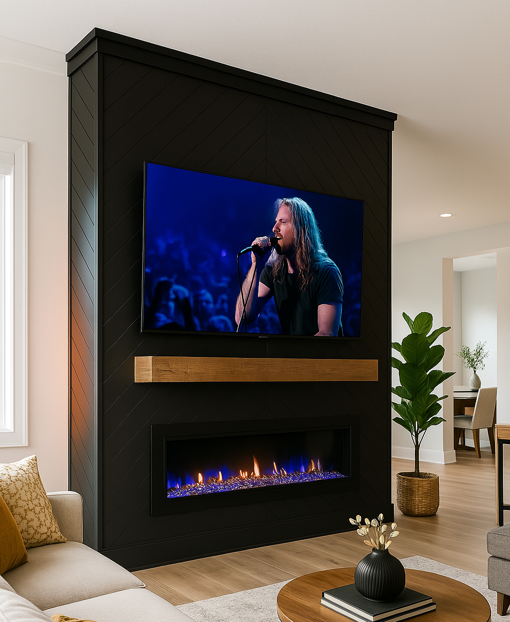 Living room with a modern black accent wall featuring a mounted flat-screen TV showing a singer. Below the TV is an electric fireplace with a blue flame. The room has a beige sofa with pillows, a plant in a woven basket, a round wooden coffee table w