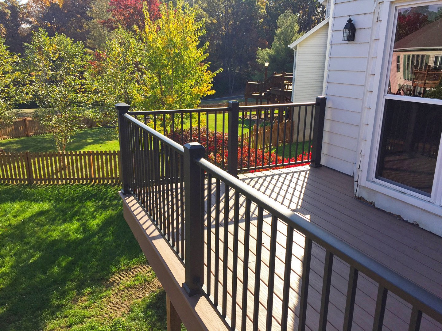 View of a wooden deck with black metal railing attached to a white house, overlooking a backyard with grass, trees, and a wooden fence, in autumn.