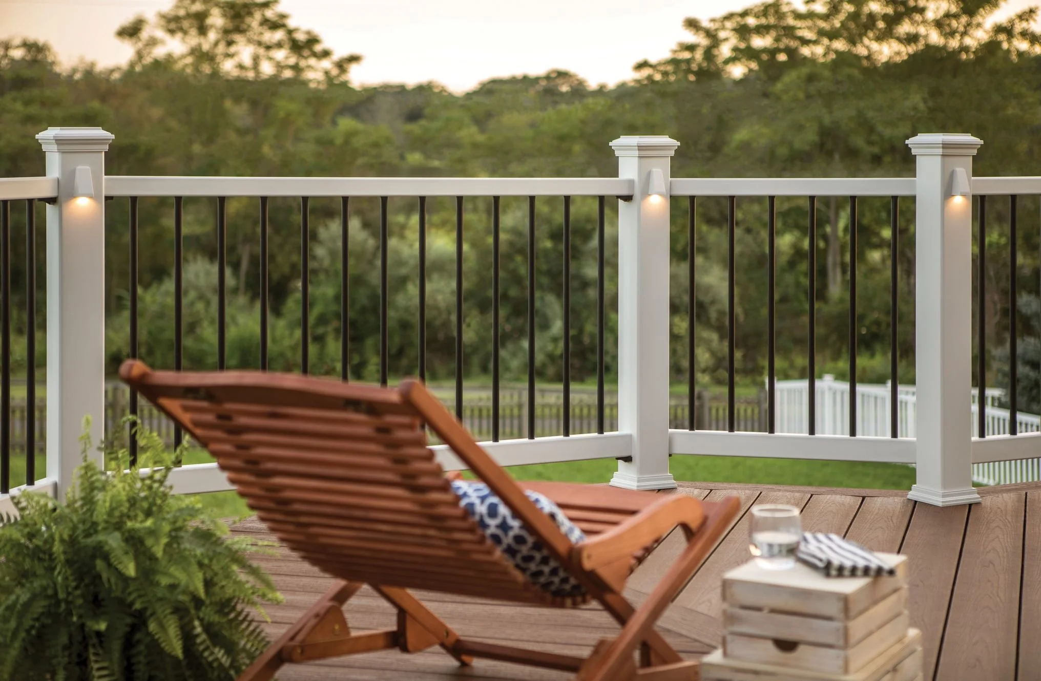 Wooden deck with a chair, a small table with a glass of water, and a potted fern, overlooking a green landscape with trees, and a white fence in the background, during sunset.