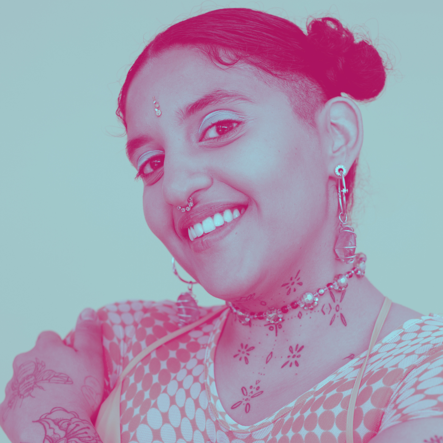 Portrait of a smiling woman with tattoos, colorful jewelry, and a short hairstyle with buns, against a plain background.
