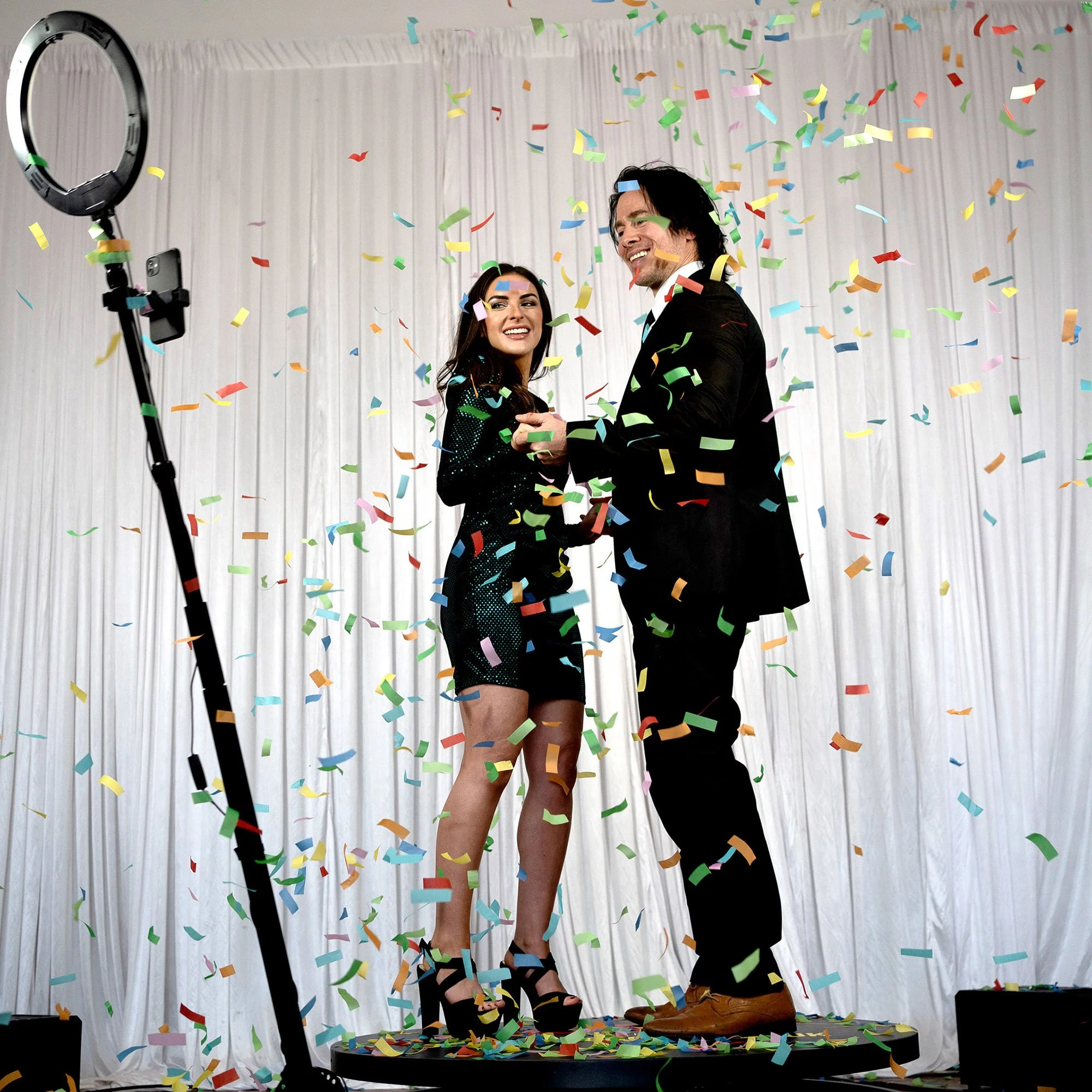 A man and woman smiling and holding hands on a stage, surrounded by colorful confetti, with a white backdrop, during a celebration or event.