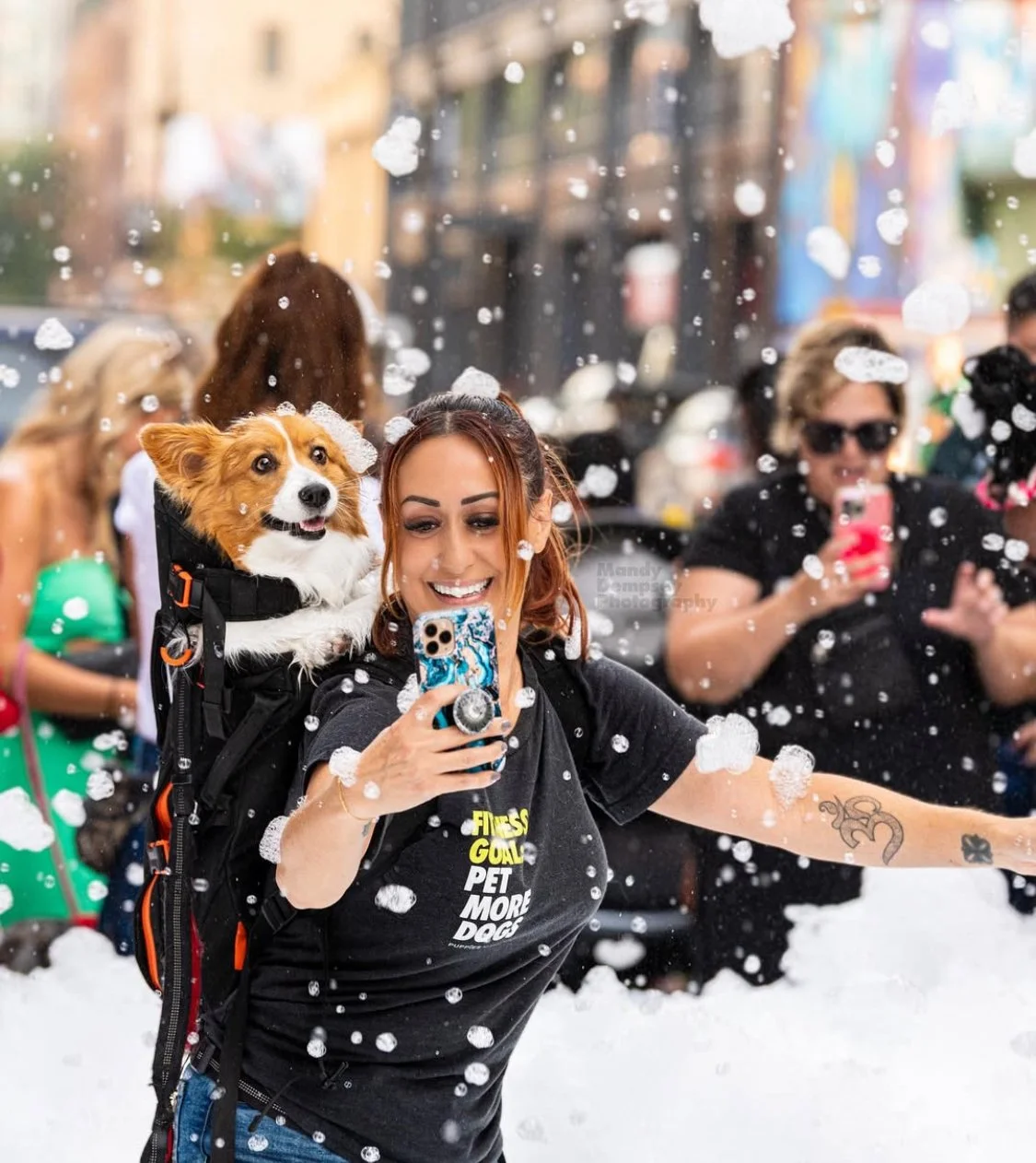 Woman with a small dog in a backpack taking a selfie during a snowstorm, with others taking photos in the background.
