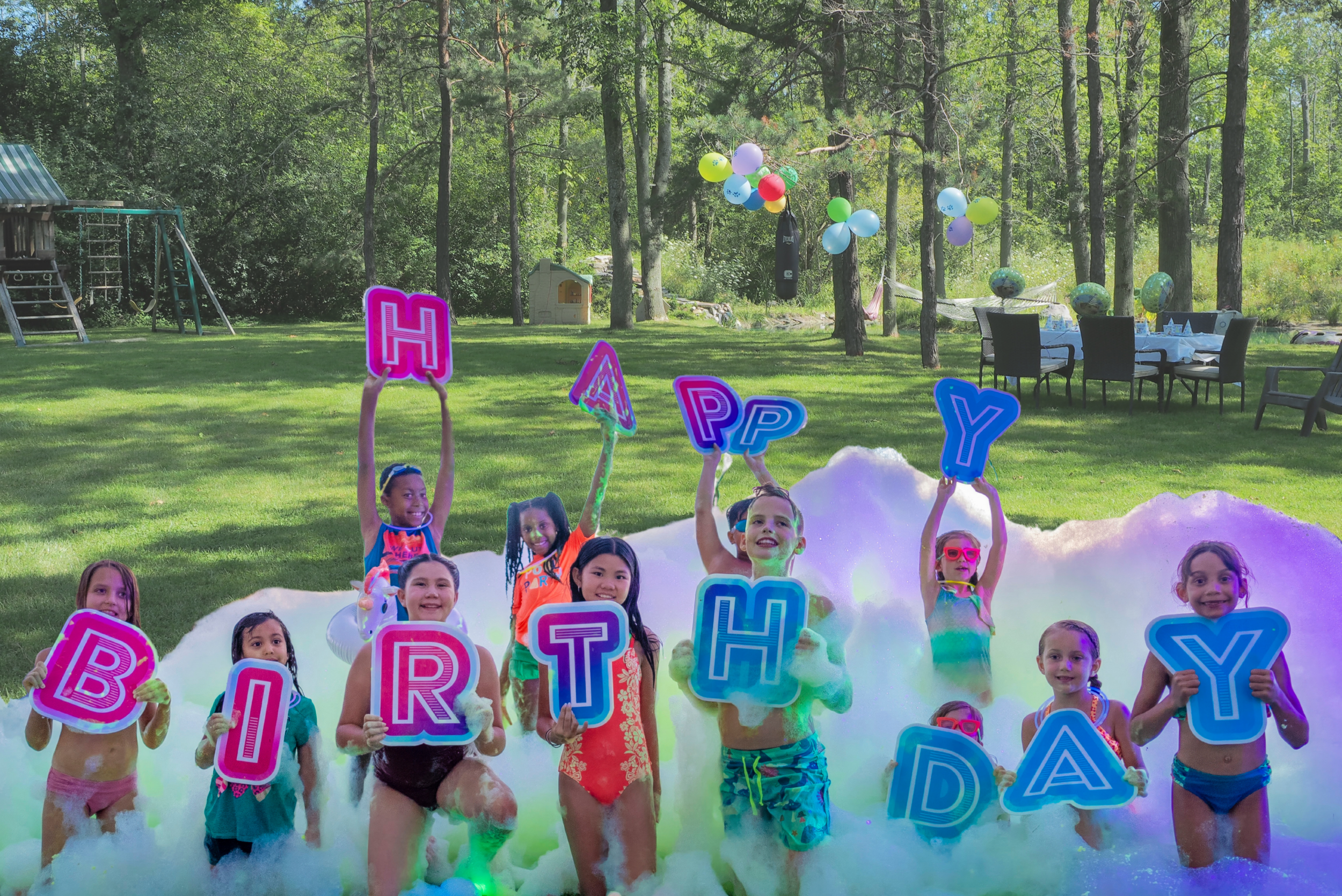 Children celebrating a birthday party outdoors in a grassy yard with foam, holding colorful letters that spell out "HAPPY BIRTHDAY." Some children are holding foam letters, others are raising the letters, and a few are wearing swimsuits. Balloons and balloons bouquets fill the air above them, and several tables and chairs are set up in the background among trees.