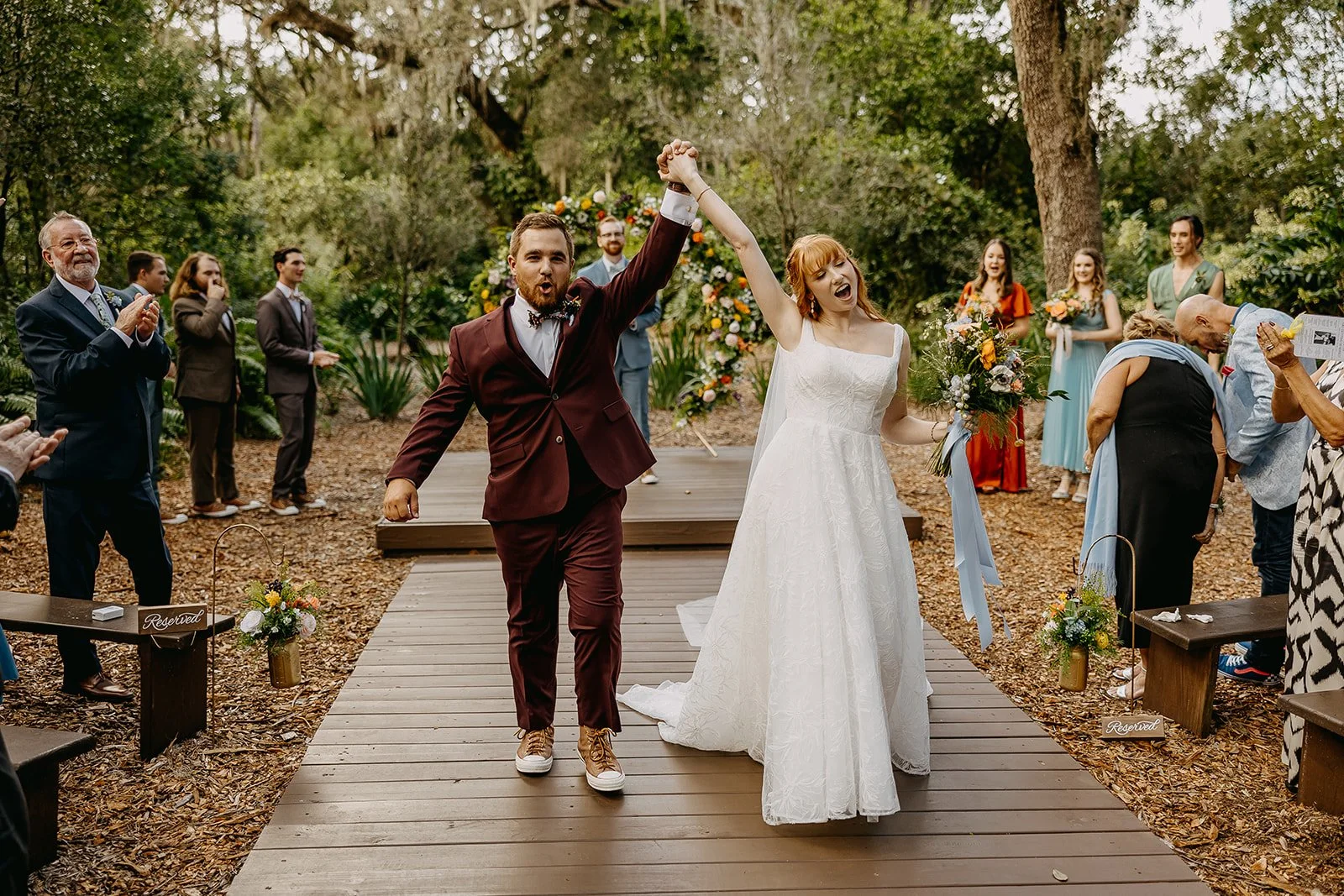 A newlywed couple walking hand in hand down an outdoor aisle, celebrating their wedding. The bride is wearing a white wedding dress and holding a bouquet, while the groom is in a burgundy suit with sneakers. Guests are clapping and cheering on both sides, with some taking photos, all amidst a natural, wooded setting.