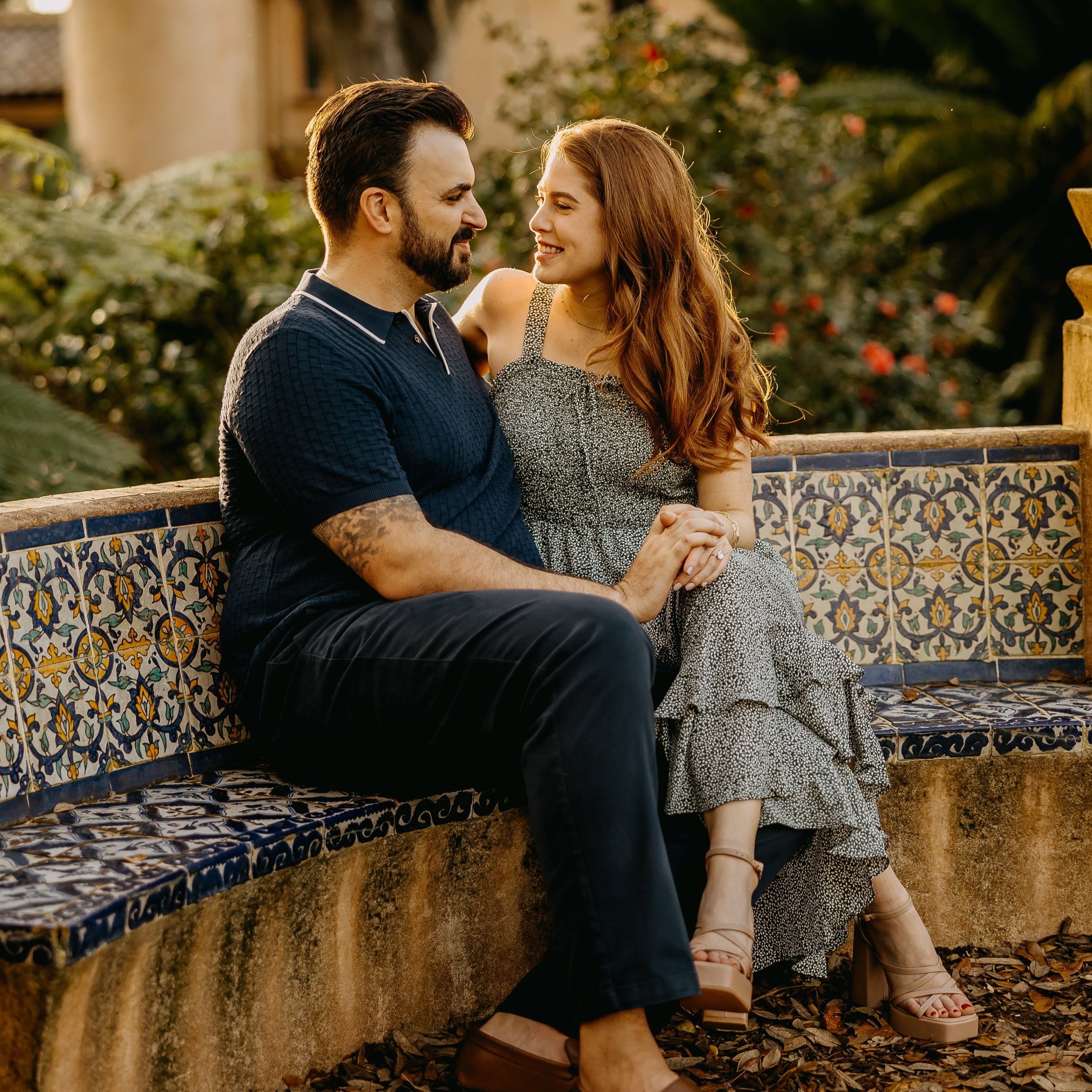A man and woman sitting close on a tiled stone bench outdoors, holding hands and smiling at each other during sunset.