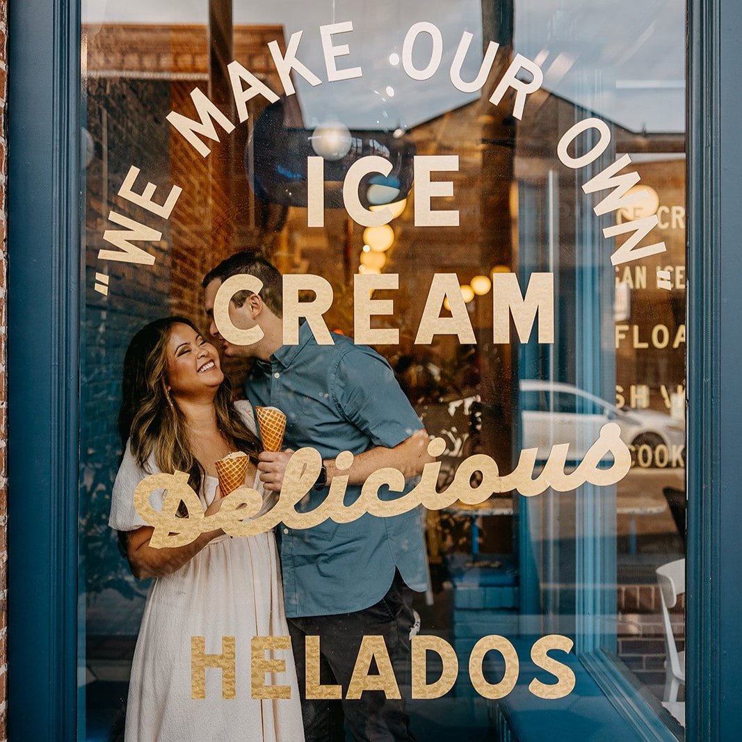 A couple inside an ice cream shop holding cones and smiling through a glass door with text advertises ice cream and desserts.