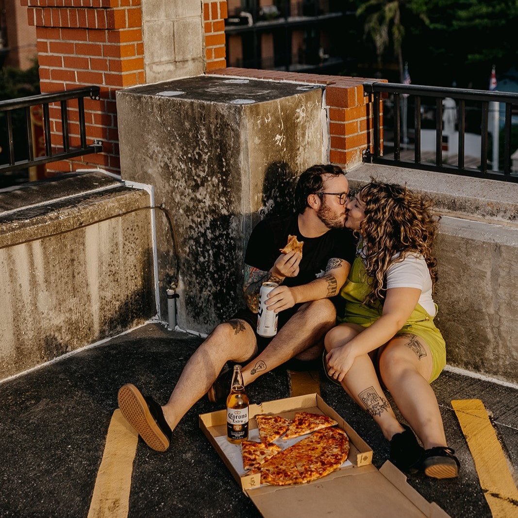 A couple sits on a parking lot rooftop at sunset, sharing a kiss, with a pizza and beer on the ground in front of them.