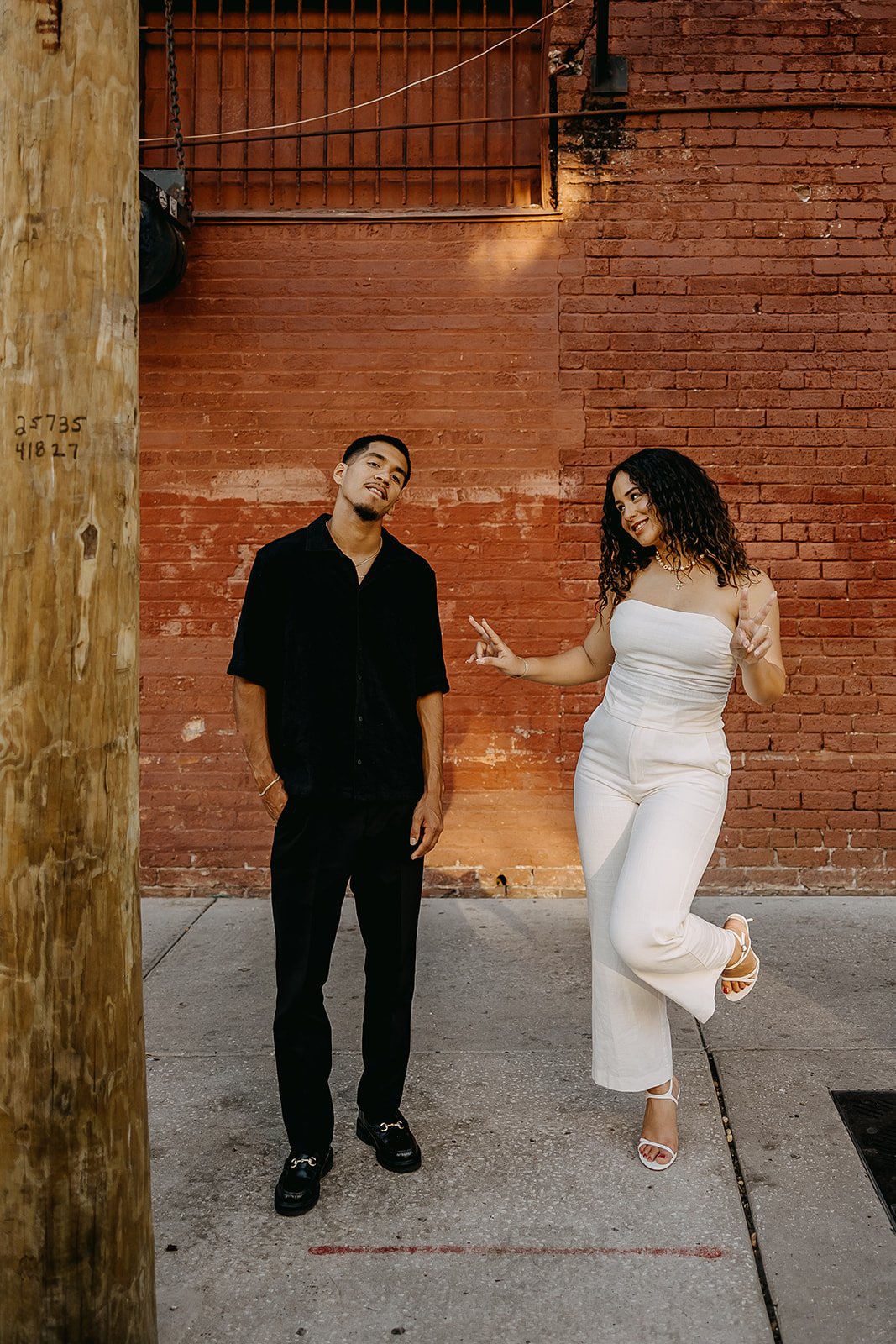 A man and woman standing on a sidewalk in front of a brick wall. The man has his hands in his pockets, dressed in black, and the woman is smiling, making peace signs with her hands, dressed in white pants and a strapless top. The man looks amused or relaxed, while the woman appears cheerful and playful.