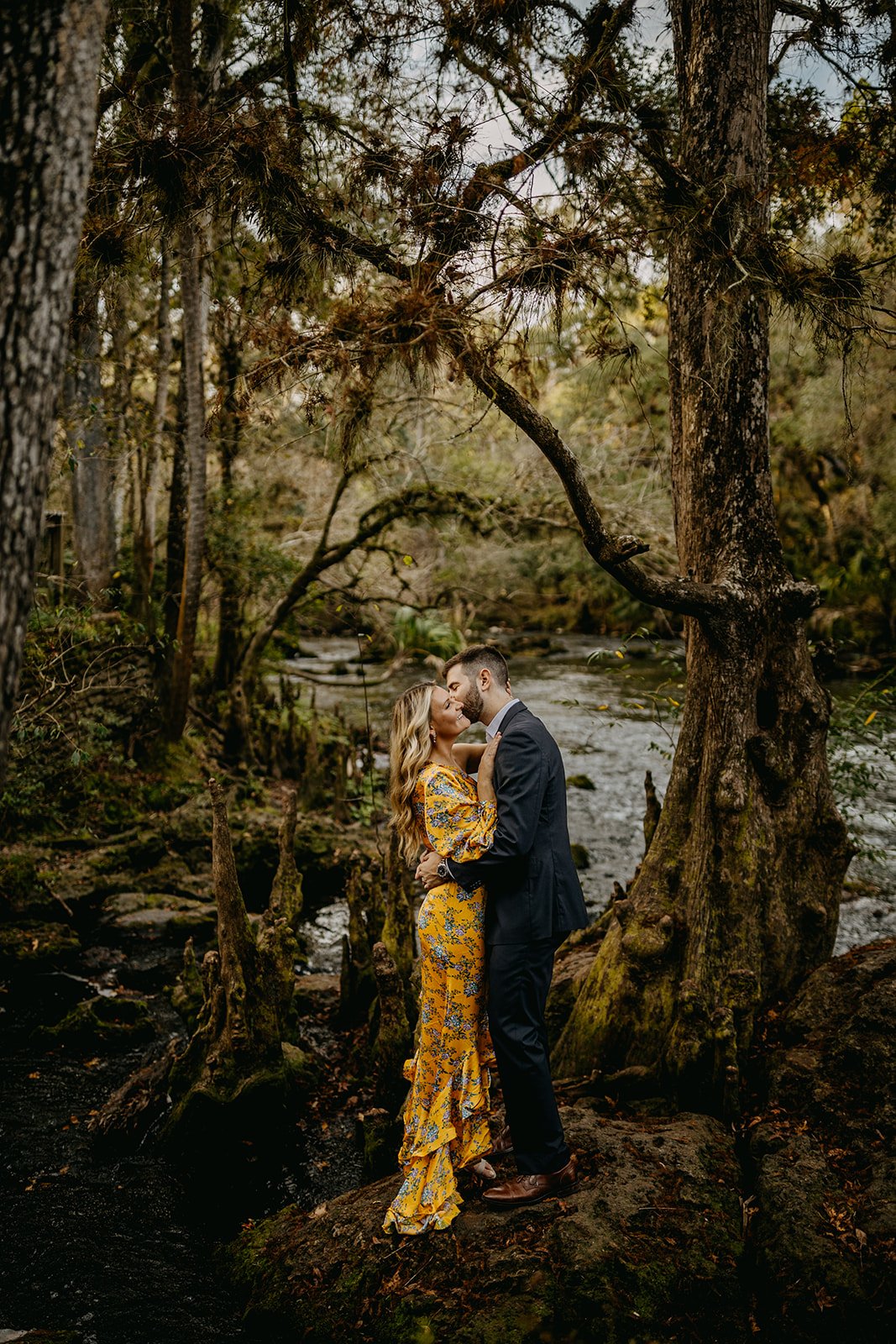 A couple embracing and kissing in a forest setting by a stream, surrounded by trees and natural scenery.