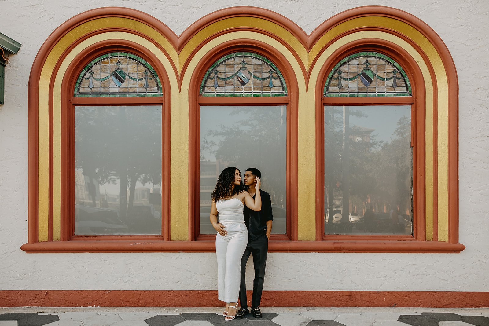 A couple standing in front of three large arched windows with stained glass details, the woman in white and the man in black, embracing each other.