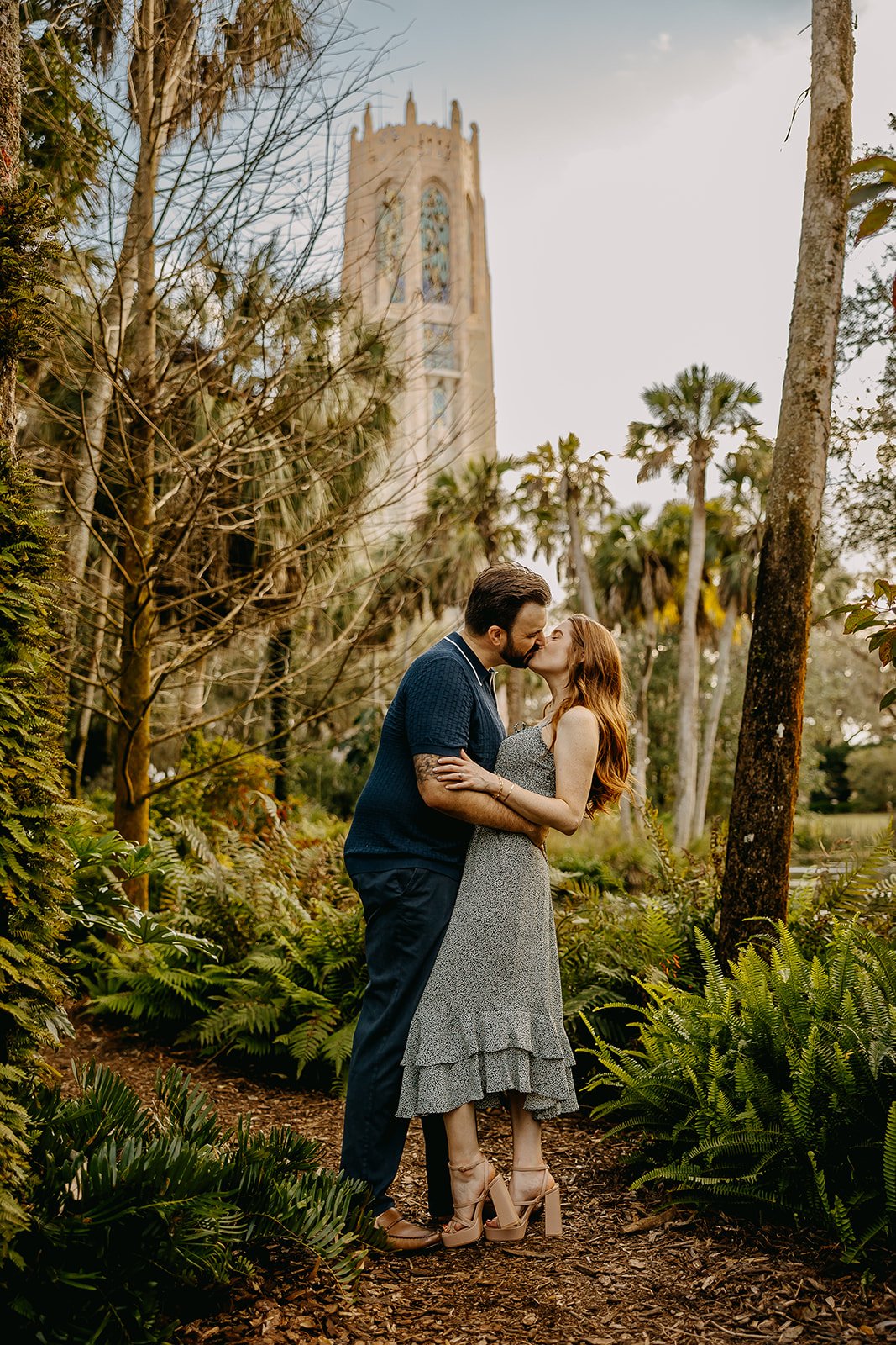 A couple kissing while standing in a lush, green garden with tall trees and ferns, with a gothic-style tower in the background.