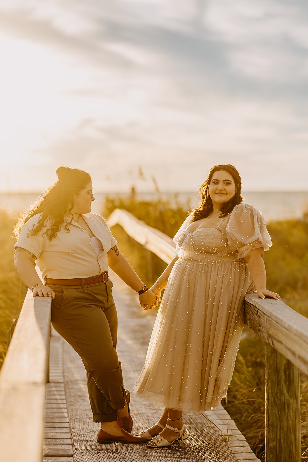 Two women holding hands on a wooden walkway near the beach during golden hour with the ocean in the background.