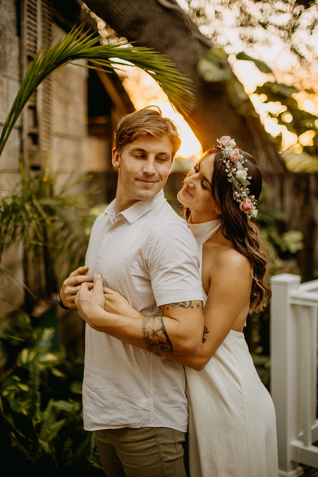 A romantic couple sharing an intimate dance outdoors at sunset, with the woman wearing a floral crown and the man with tattoos on his arm, surrounded by lush greenery and a rustic wooden structure.