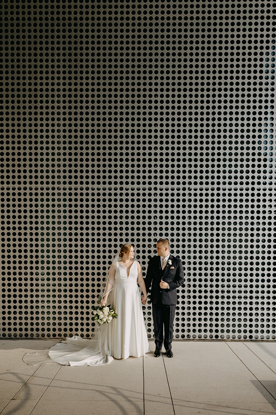 A bride and groom standing hand in hand outdoors at night in front of a large black perforated metal wall. The bride is wearing a white gown with a v-neckline and holding a bouquet of white flowers. The groom is dressed in a black suit with a boutonniere, looking at the bride.