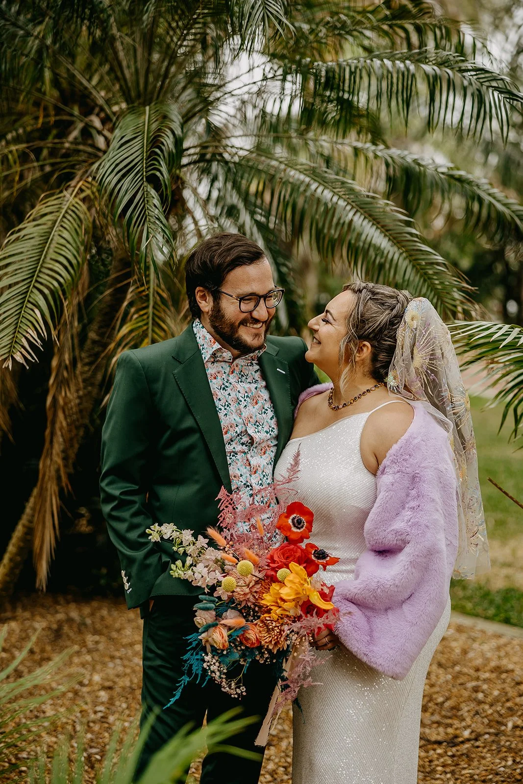 A bride and groom on their wedding day, smiling at each other outdoors with palm trees in the background. The bride wears a white dress, lavender faux fur shawl, and veil, holding a colorful bouquet. The groom wears a dark green jacket and glasses.