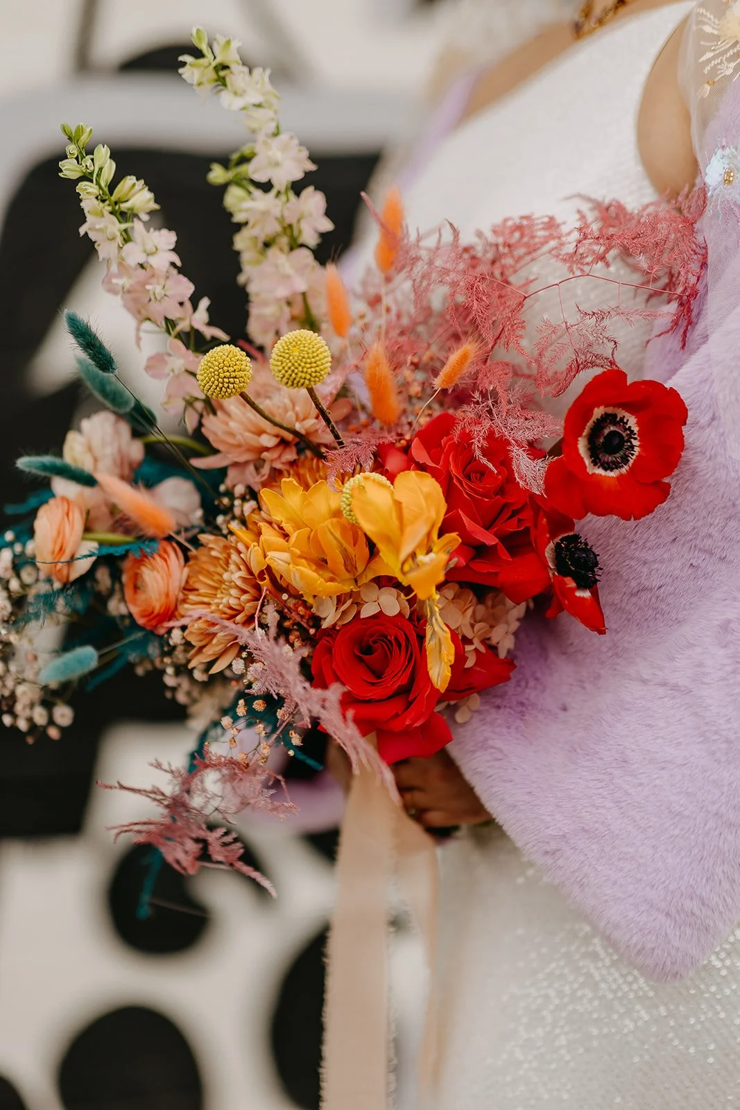 A person wearing a pink fuzzy sweater holding a colorful bouquet of flowers featuring red roses, yellow and peach flowers, white, pink, and teal accents, with various textured green and pink foliage.