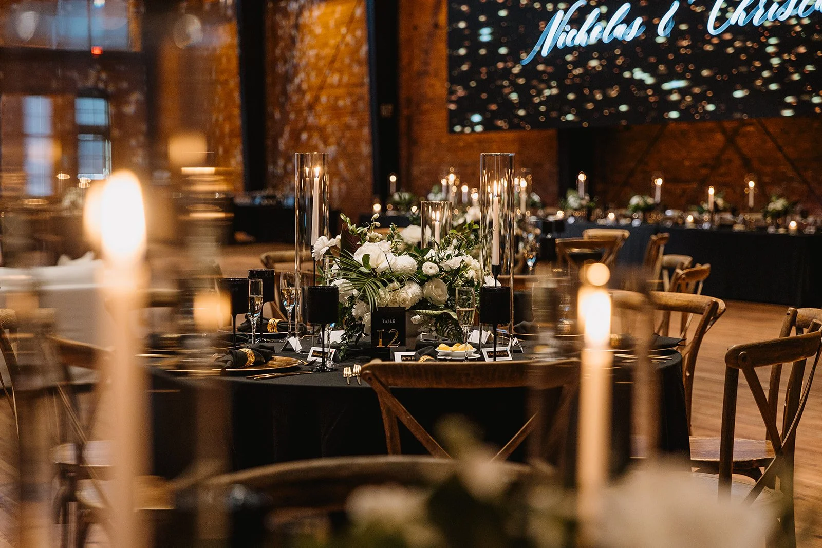 Elegant wedding or event dining setup with black tablecloths, floral centerpieces with white flowers, candles, and decorative tall glass vases in a brick-walled venue.