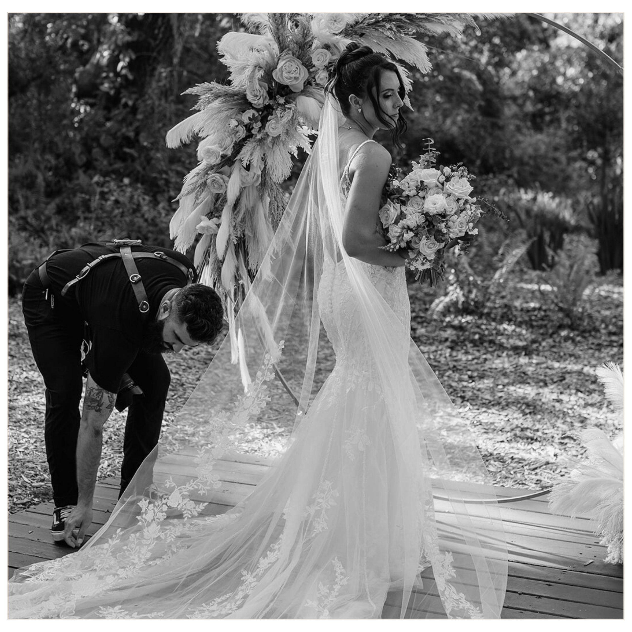 A bride in a wedding dress with a long, flowing veil is holding a bouquet of flowers in an outdoor setting. A person is adjusting the train of her dress.