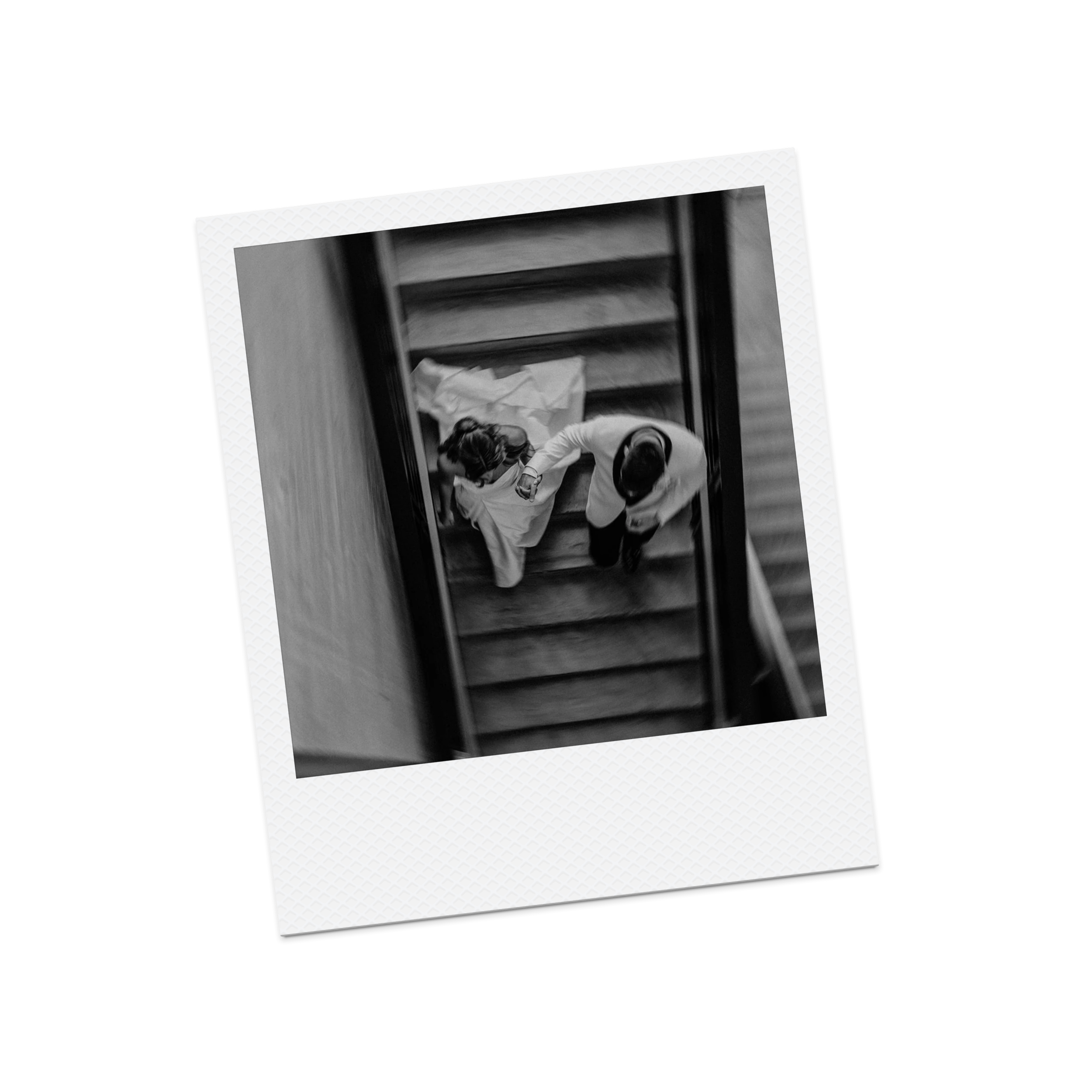 A black and white photograph of two people on a staircase, one seated and the other standing, taken from above. The seated person appears to be a woman with long hair, and the standing person is a man in a white shirt.