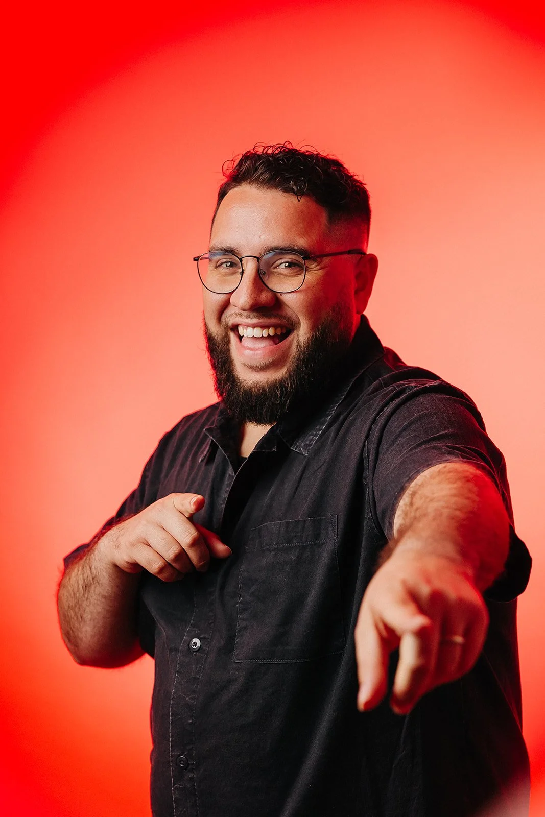 A man with glasses, a beard, and a joyful expression pointing towards the camera, standing against a red gradient background.