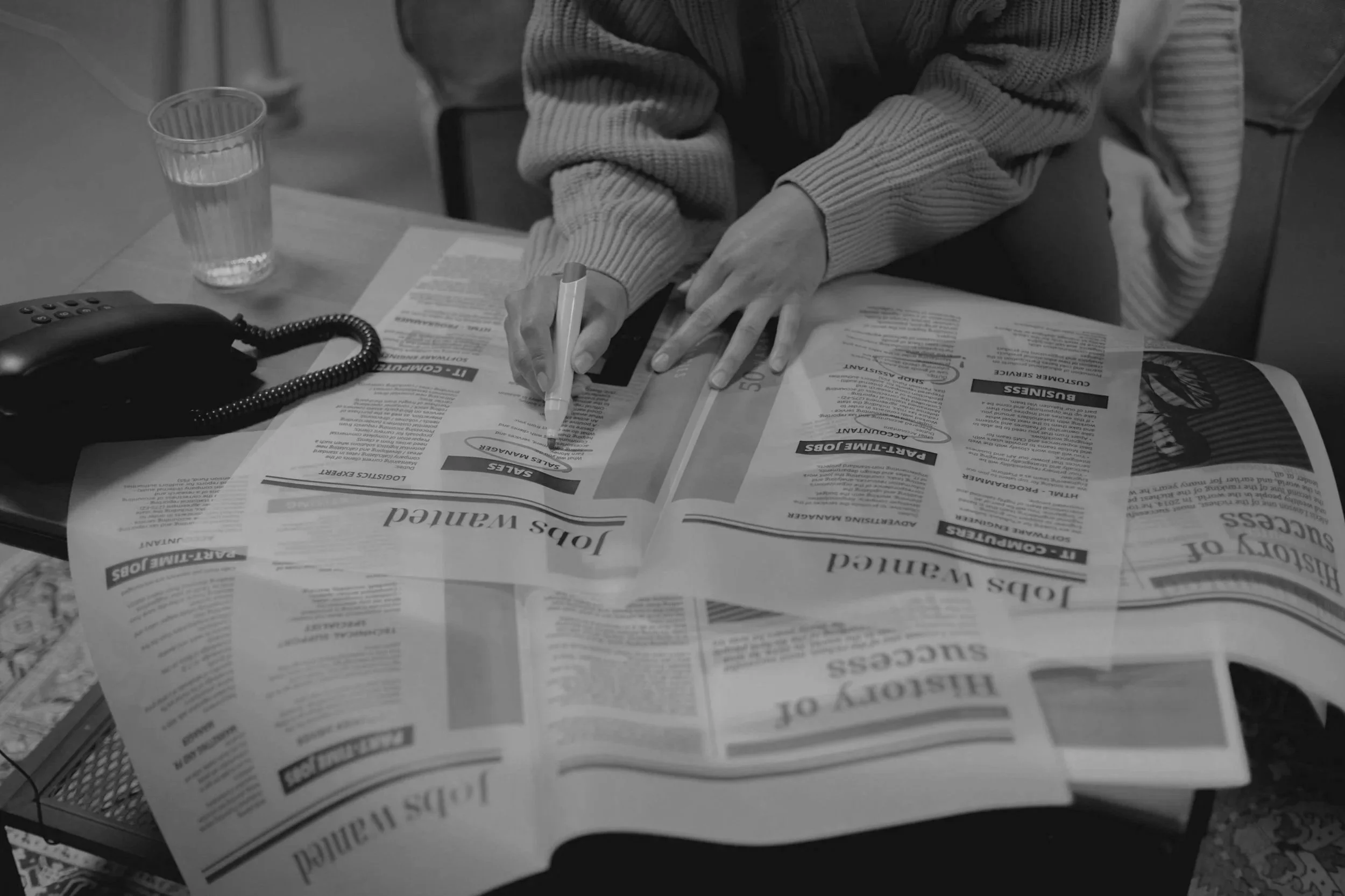 A person in a sweater writing on a newspaper with a pen in a busy setting. There is a glass of water and a telephone on the table.