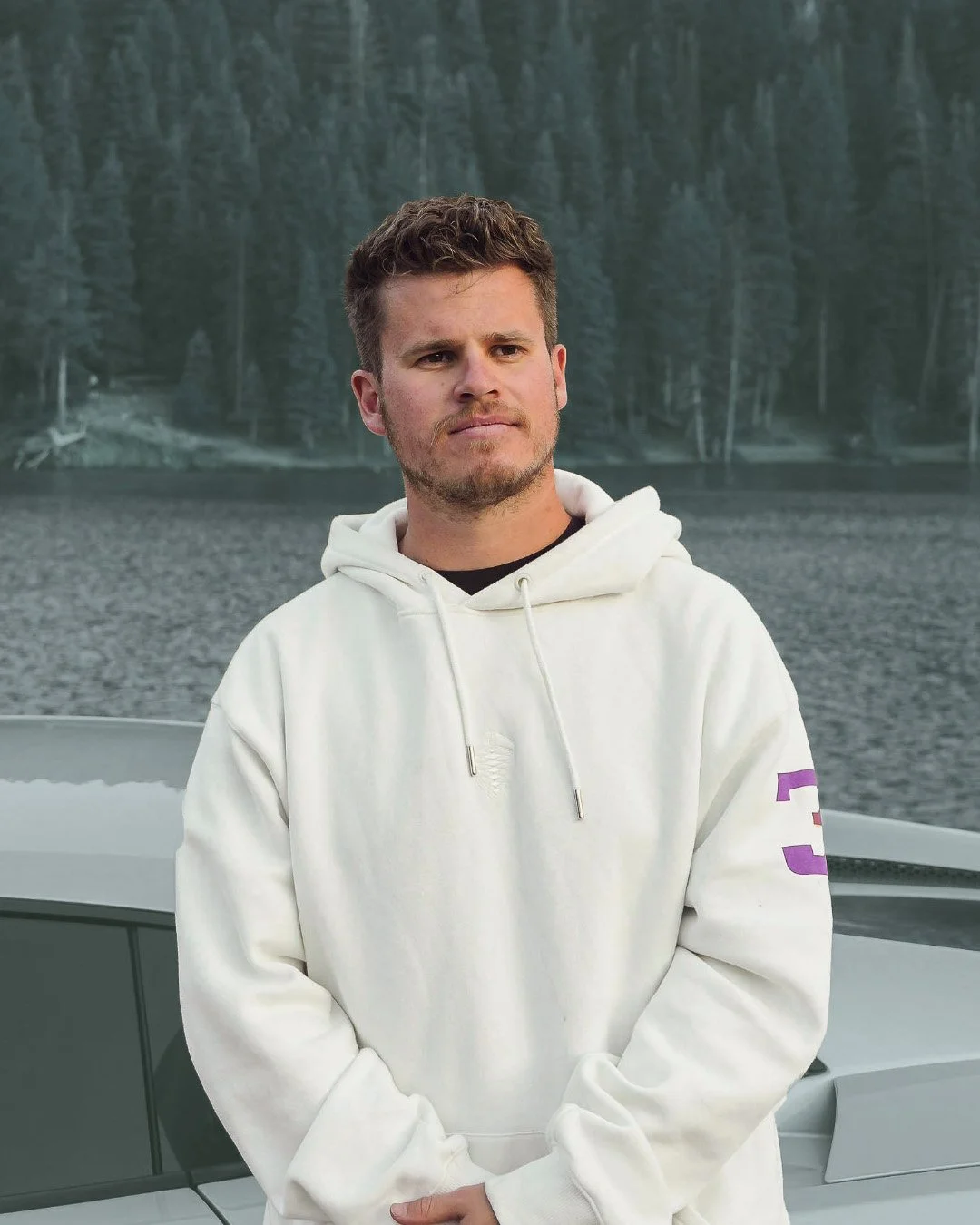 Man in a white hoodie standing outdoors near a car with a body of water and forest in the background.