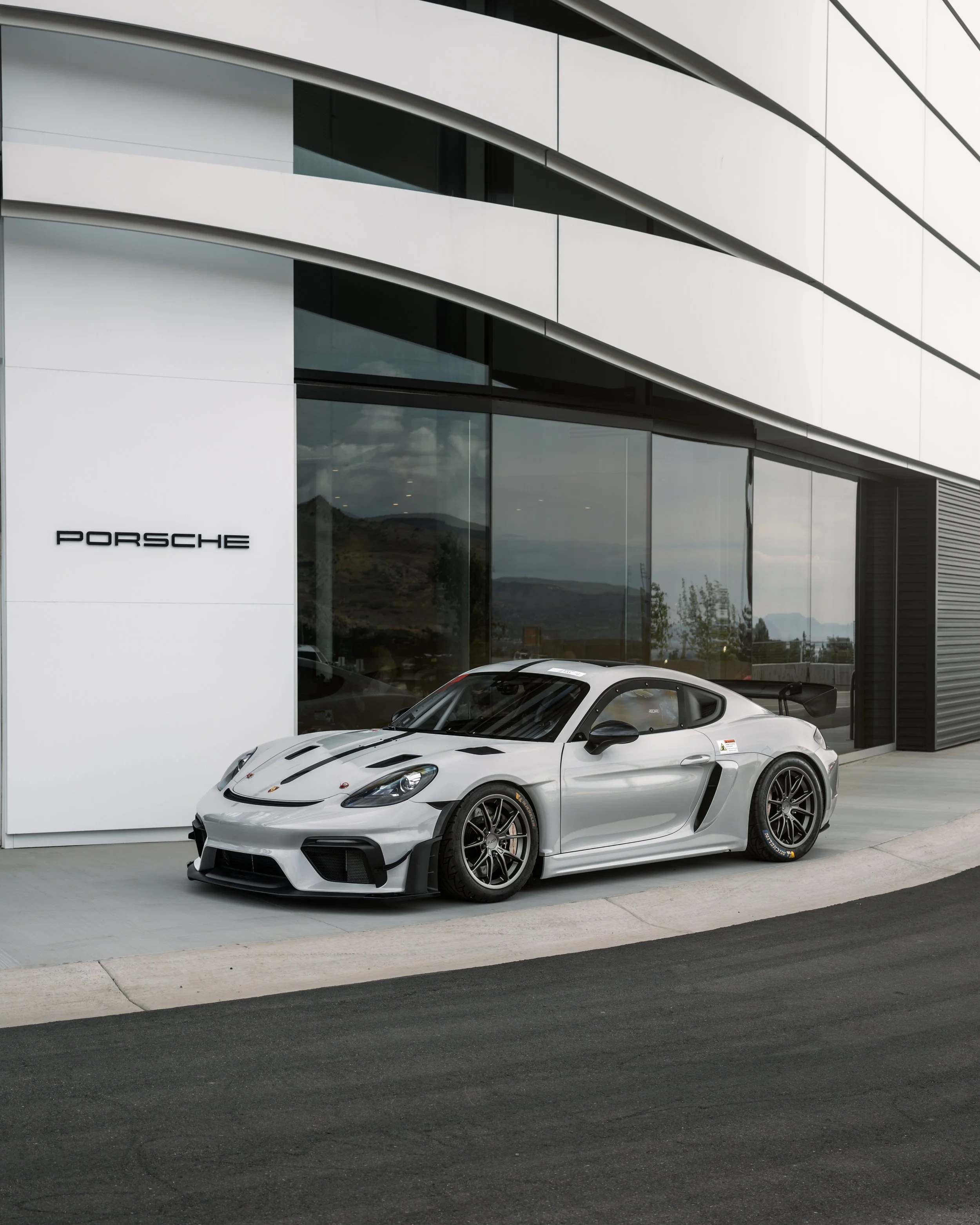 A silver Porsche sports car parked outside a modern building with large glass windows and white paneling.