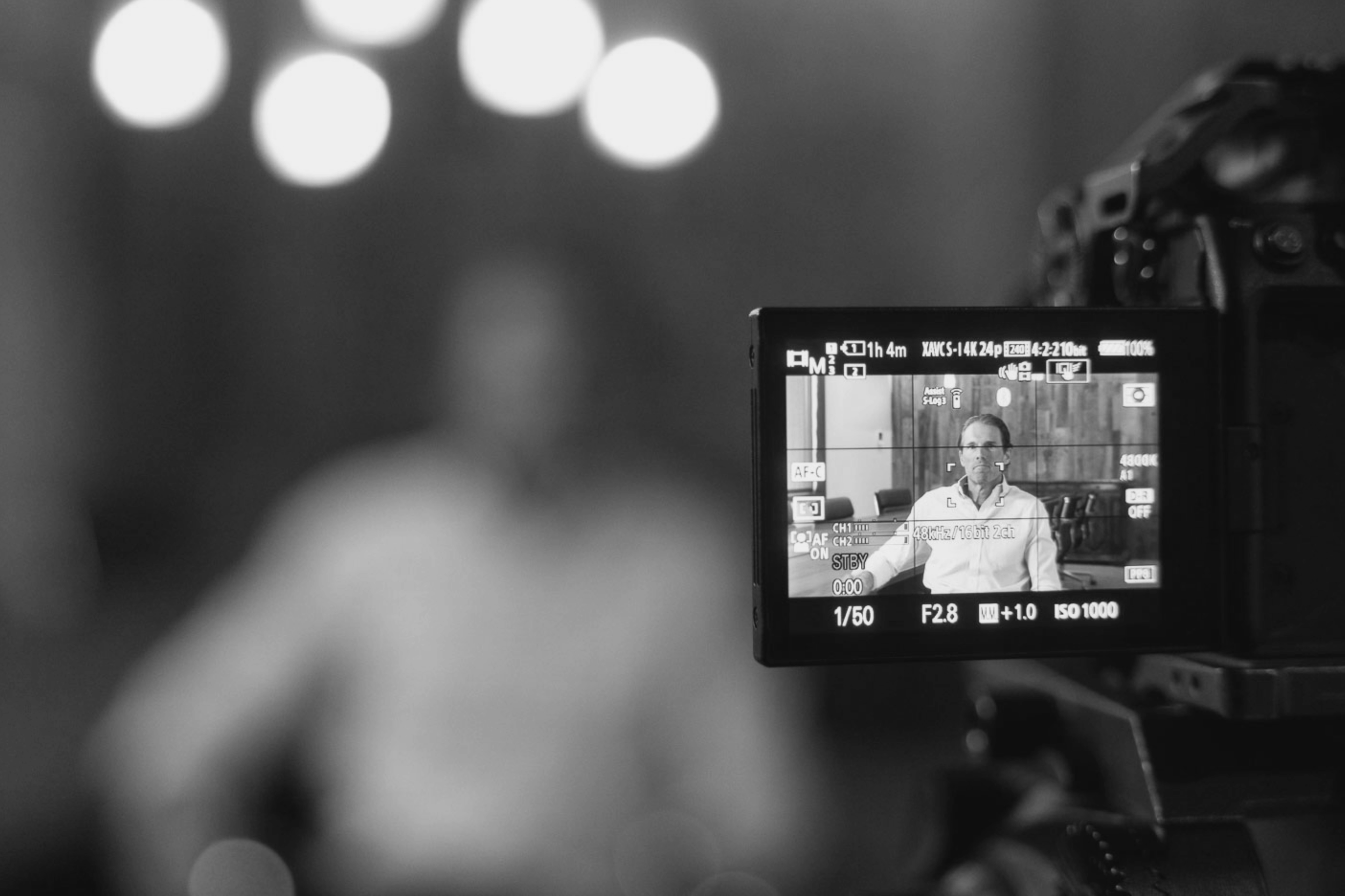 A camera's LCD screen showing a man seated in front of a camera, being recorded for a video or photo shoot, with various camera settings visible.