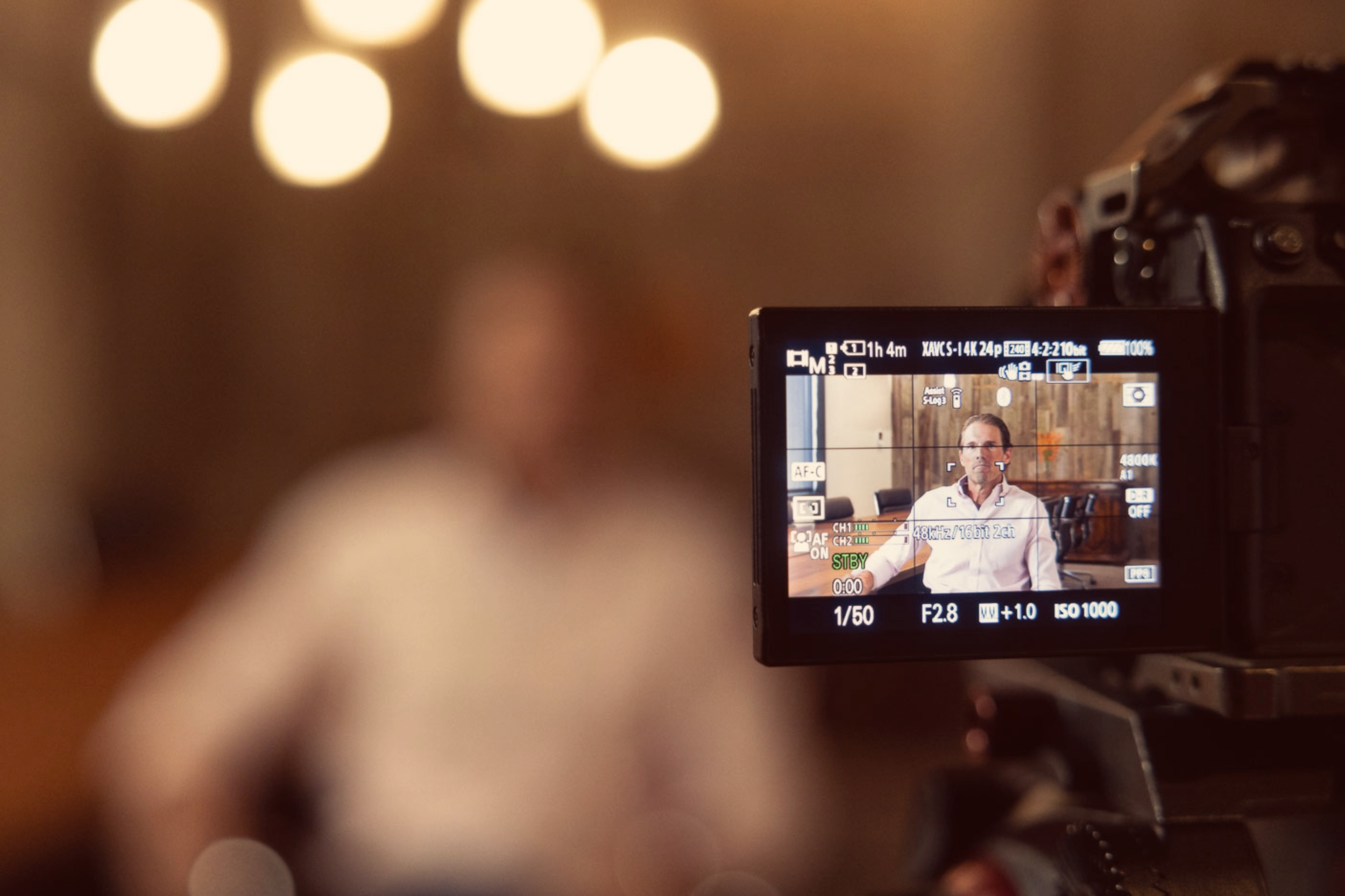 A digital camera screen showing a man in a white shirt sitting in a room with wooden walls, being recorded for a video or interview.