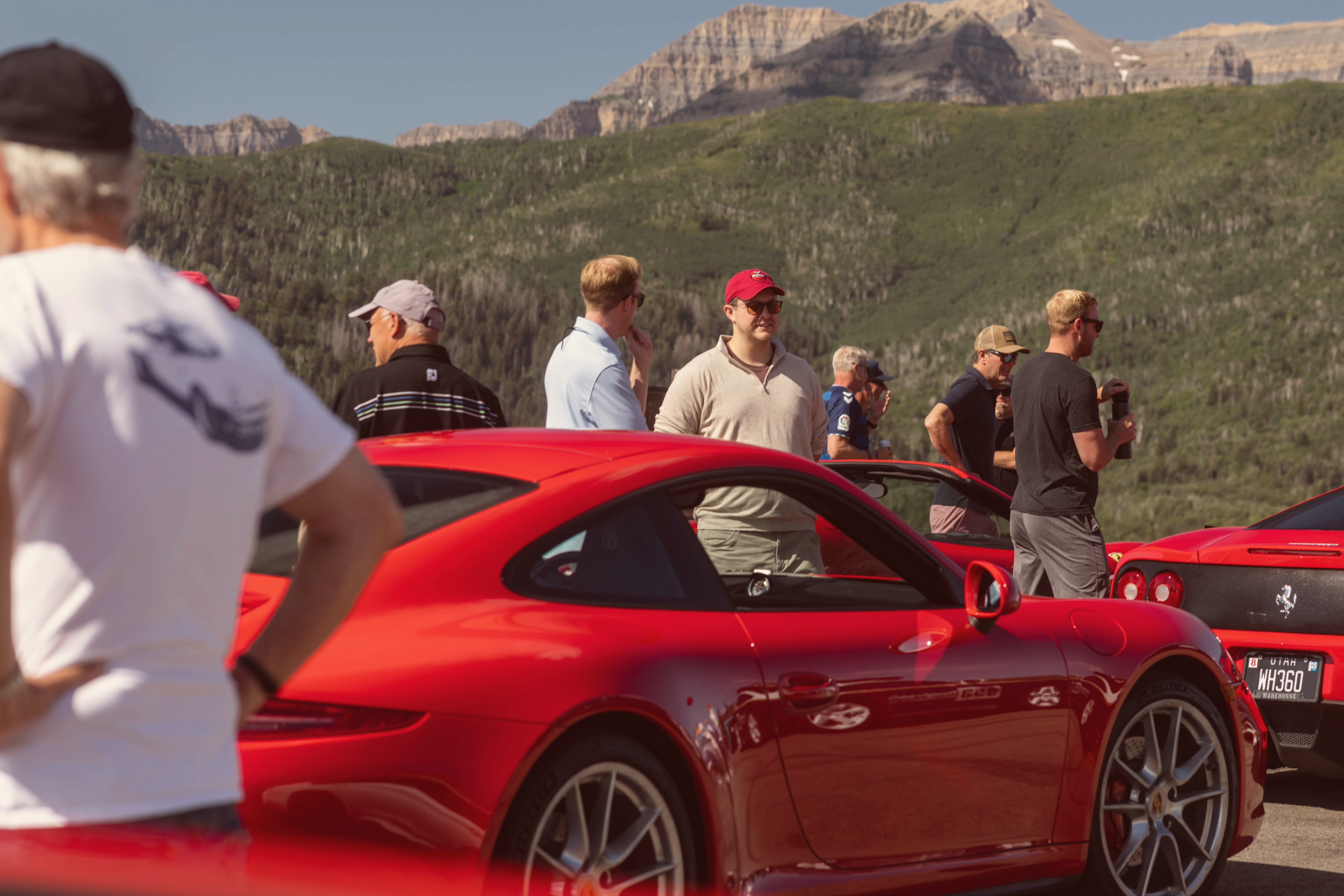 Group of people standing next to red sports cars in a scenic outdoor setting with mountains in the background.