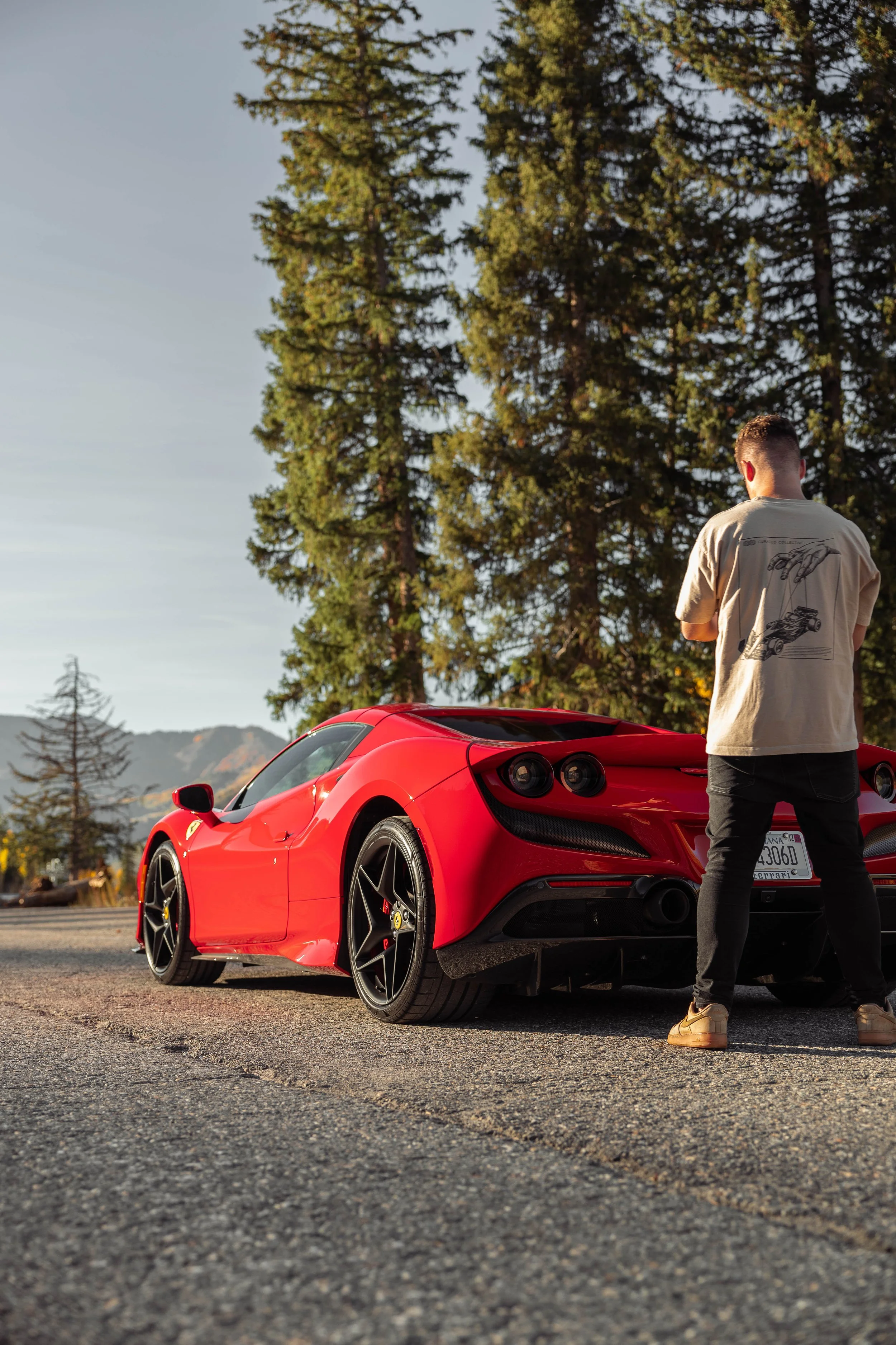 A Ferrari F8 parked on the side of the road with a man standing nearby. The background features tall trees and a mountain landscape.