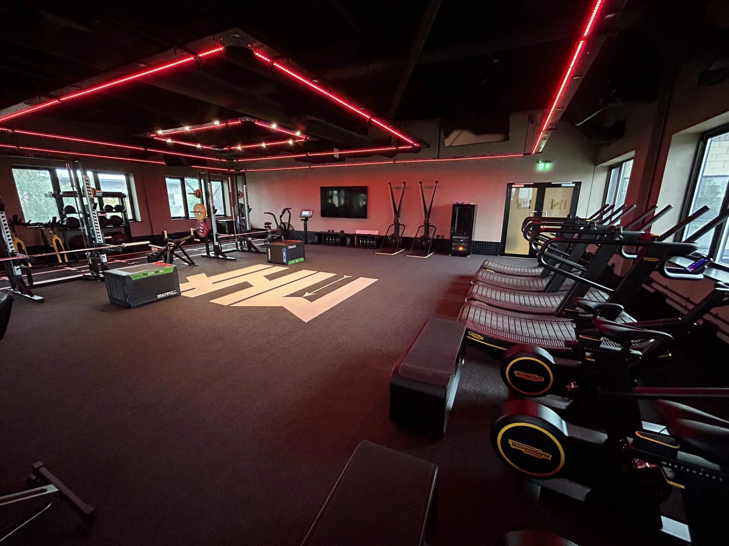 A modern gym with workout equipment and black cardio machines, illuminated by red LED ceiling lights.